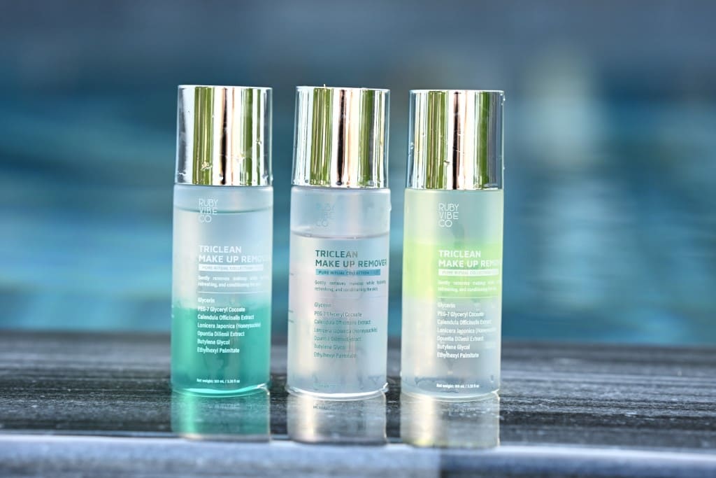 Three bottles of Ruby Vibe Co Triclean Make Up Remover in blue, clear, and green variations, lined up on a tiled surface next to a pool