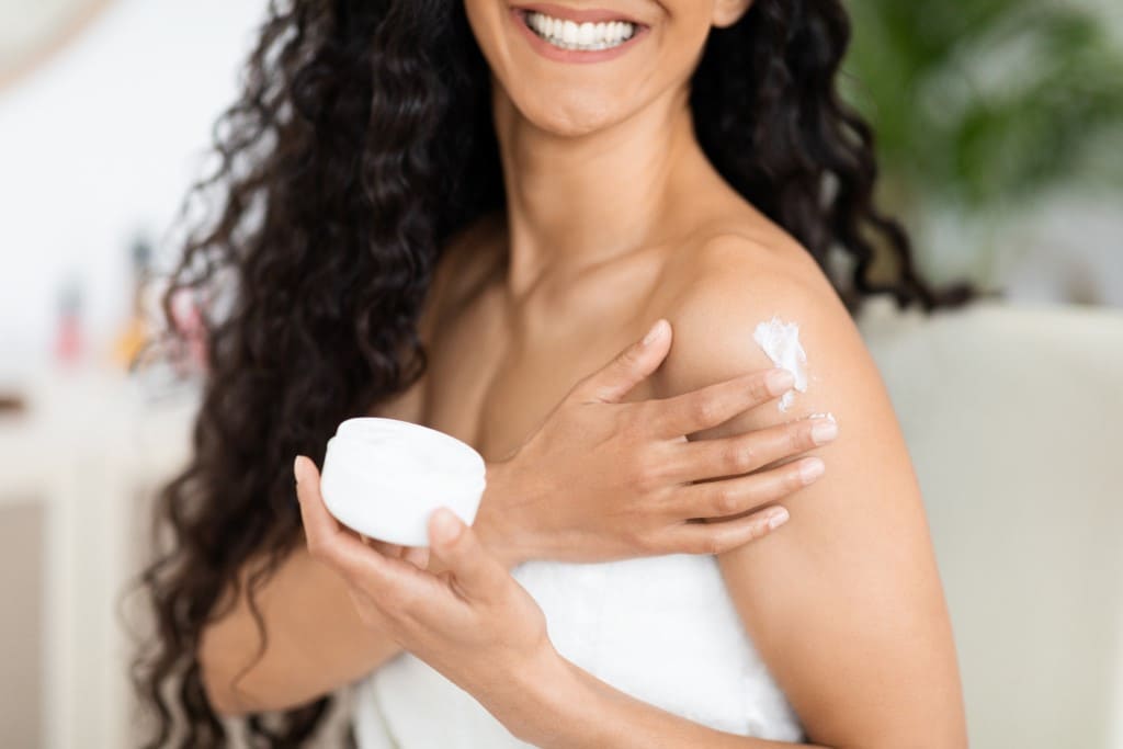 A woman applying body lotion to her shoulder.