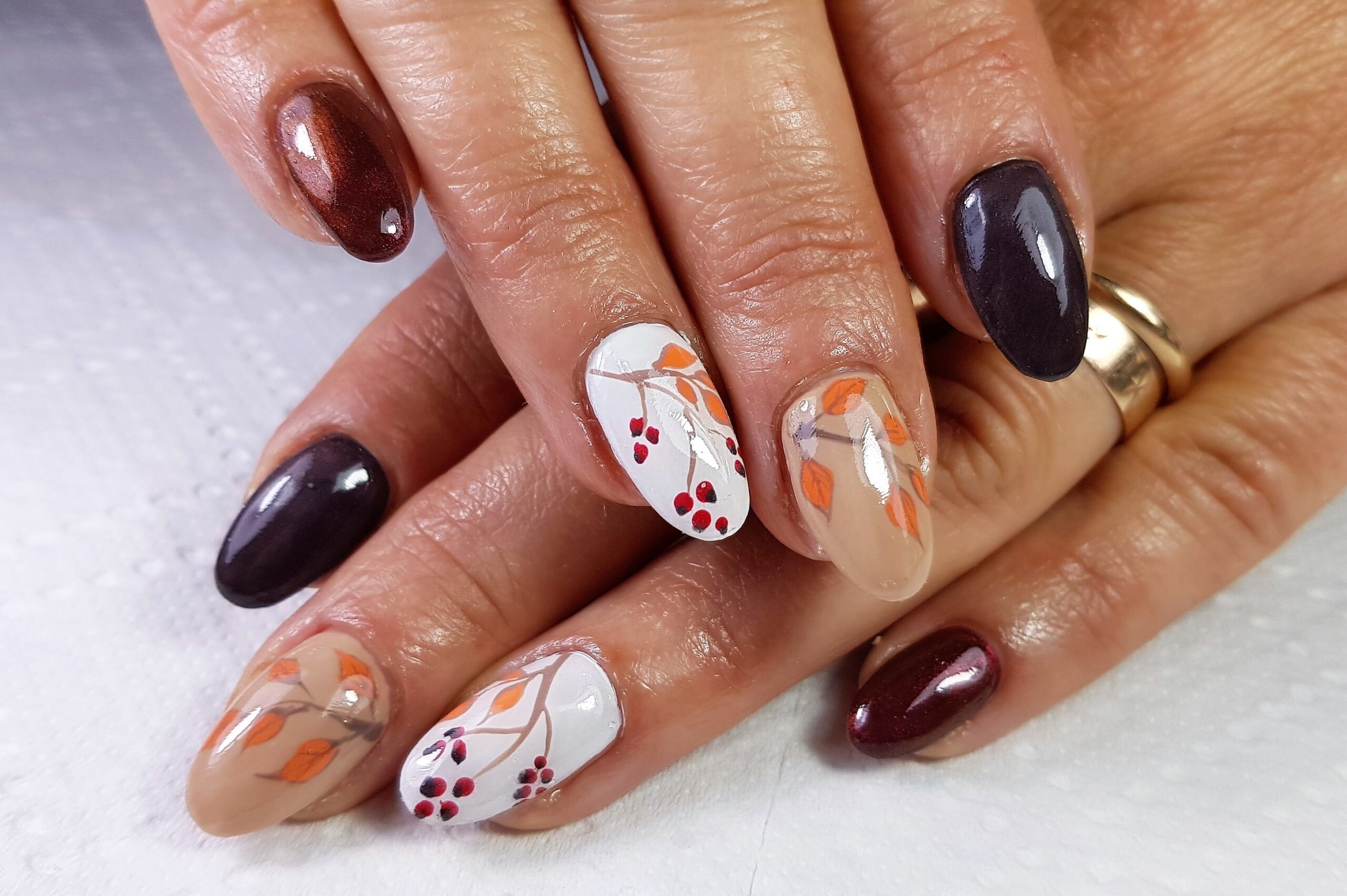 A close-up of a person's hands with a cozy and chic autumn manicure. The nails feature a mix of colors, including deep plum, rich brown, and designs with hand-painted autumn leaves and berries on a white and beige base, perfect for fall nail inspiration.