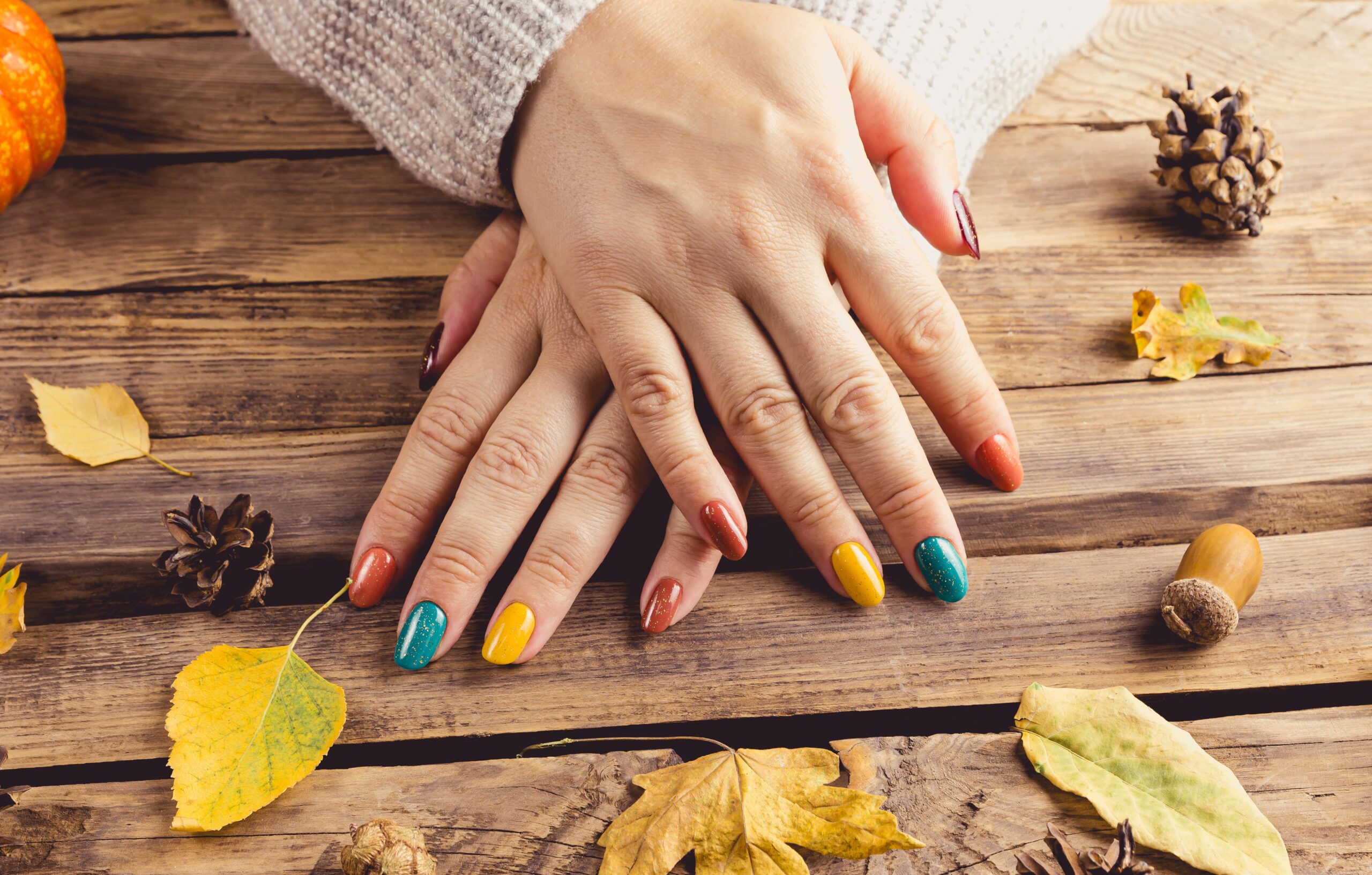 A person's hands with a multi-colored nail design rest on a rustic wooden table decorated with autumn leaves and pinecones. The nails are painted in fall shades of orange, yellow, and teal with a glitter finish, providing a guide to creating a cozy, chic manicure for the season.