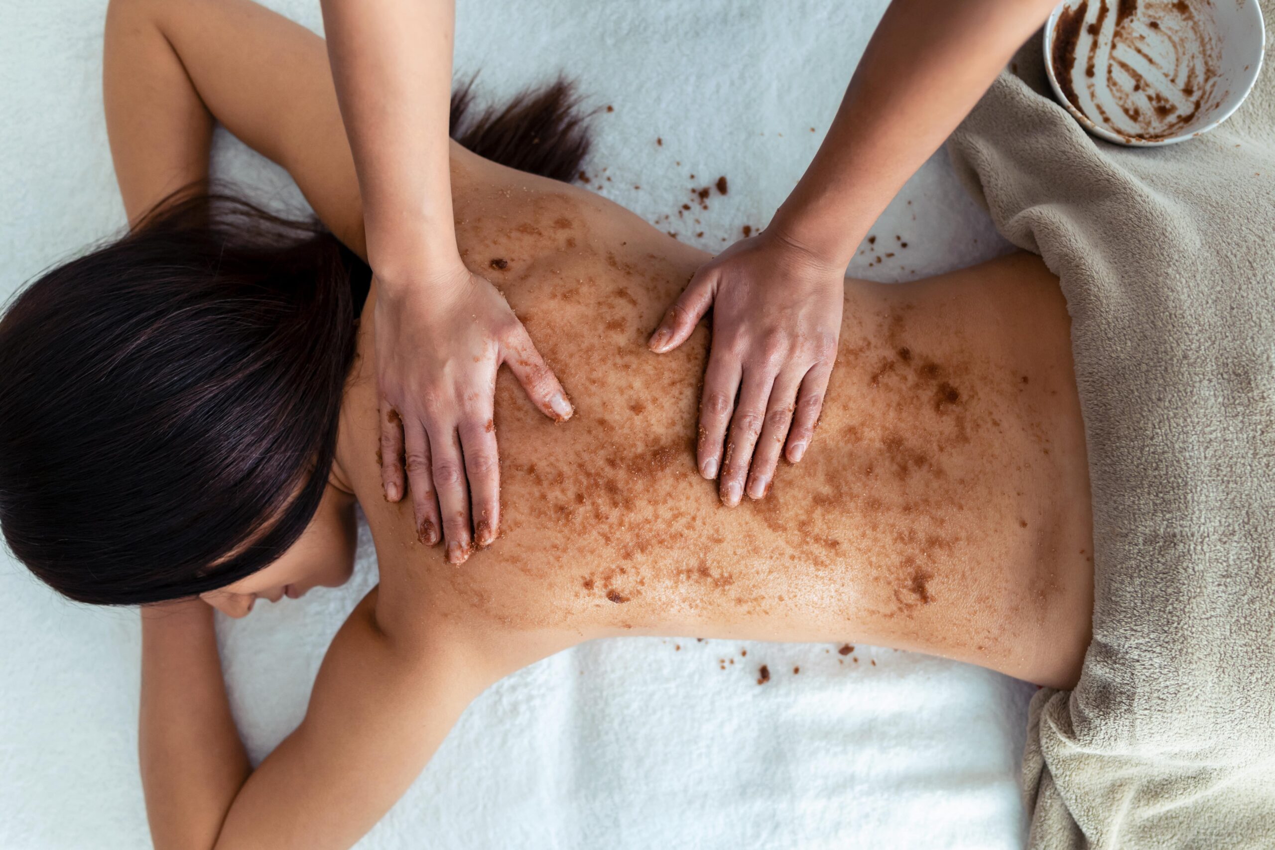 A professional applies a gritty scrub to a person's back, representing the type of physical exfoliation that should be avoided in a back acne treatment routine to prevent irritation.