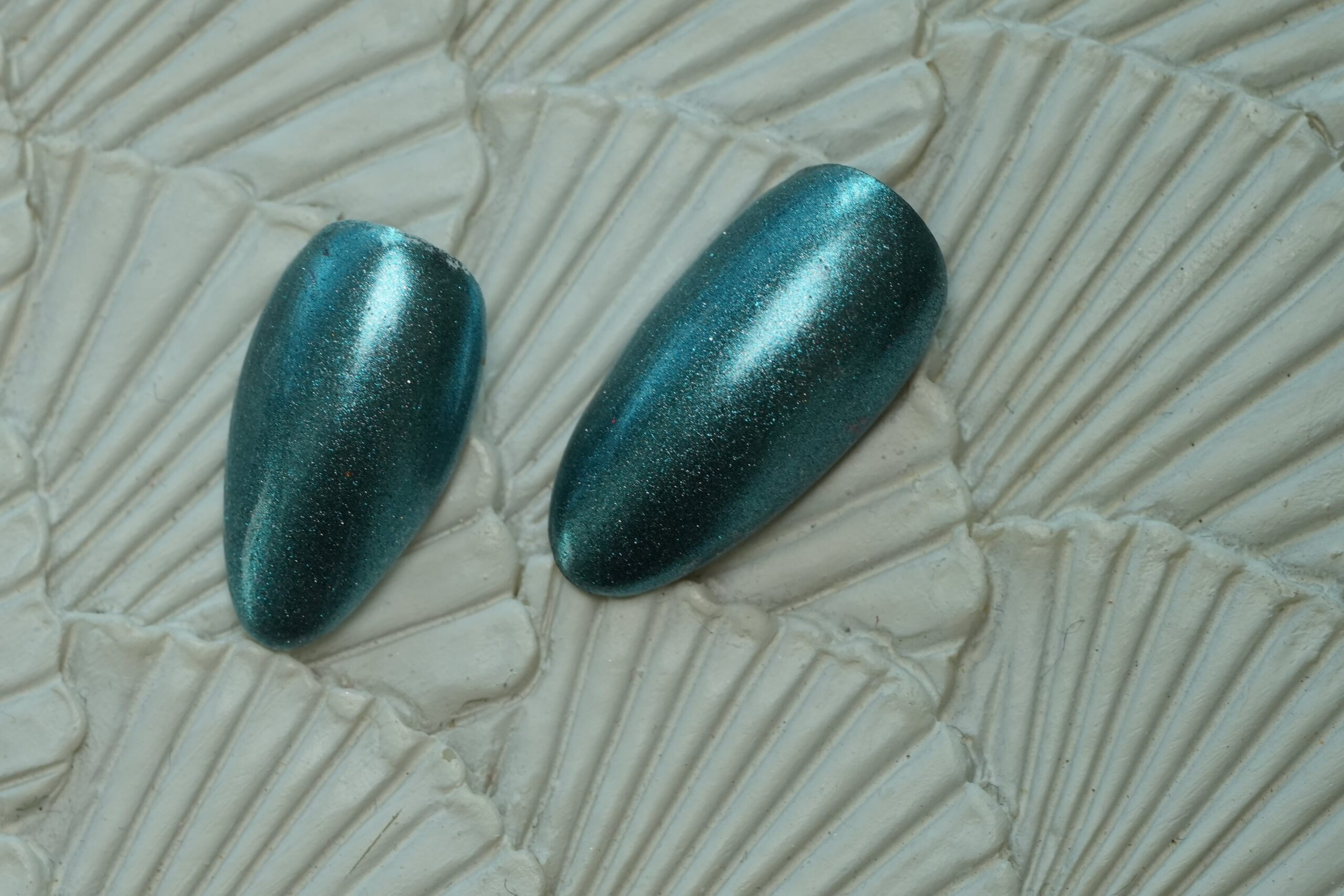A close-up of two almond-shaped nail tips with a vibrant teal chrome finish, highlighted by fine silver glitter particles for a sparkling effect.