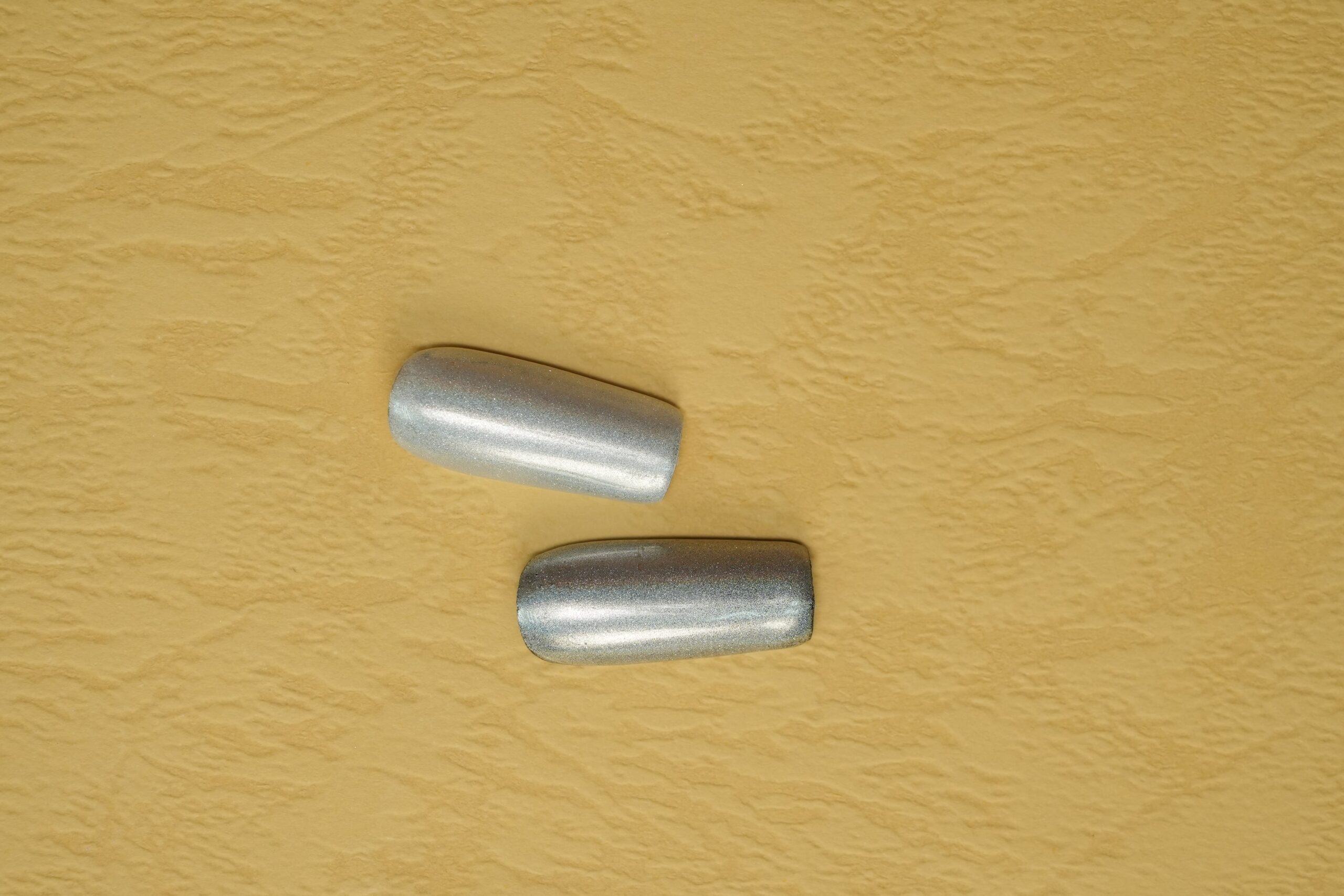 Two short, rectangular nail tips with a shimmering silver chrome powder finish, set against a textured gold background to emphasize the metallic shine.
