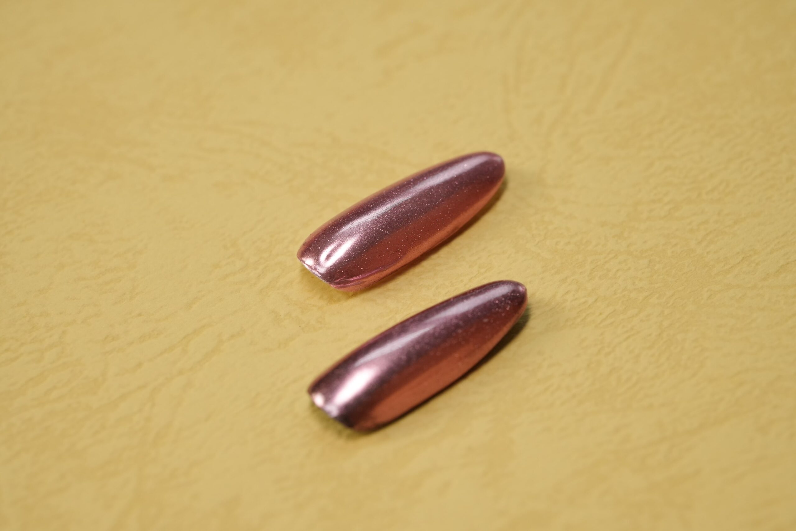 A pair of long, almond-shaped nail tips featuring a trendy rose gold chrome finish, showing a metallic and reflective surface.