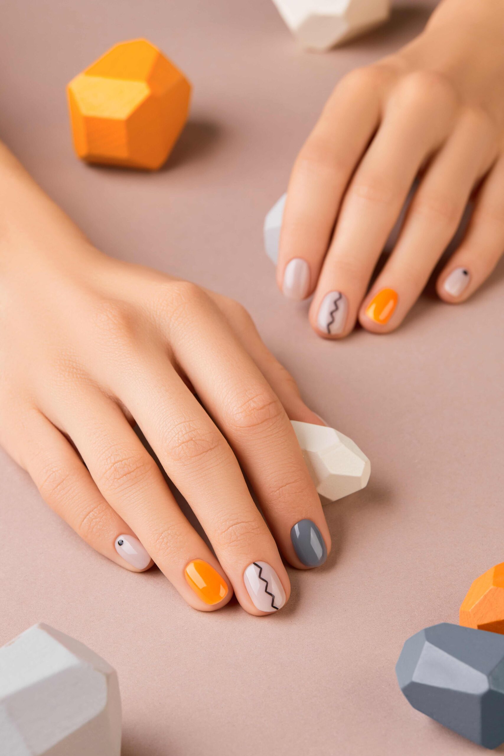 A person's hands with a chic, minimalist nail art design. The nails are painted in a cozy fall palette of grey, nude, and orange, with simple zig-zag lines and dots, providing an easy guide for creating a stylish and modern DIY fall manicure.