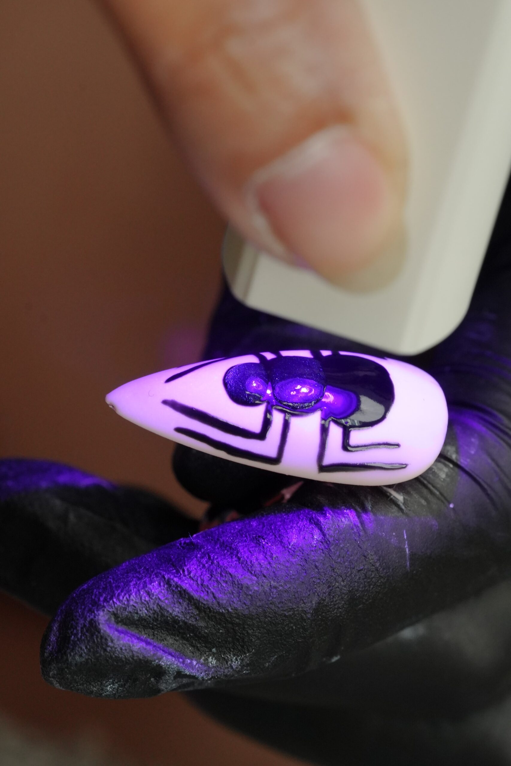 A person uses a UV light to cure a nail with a hand-painted black spider design, a key step in a DIY Halloween nail art tutorial.