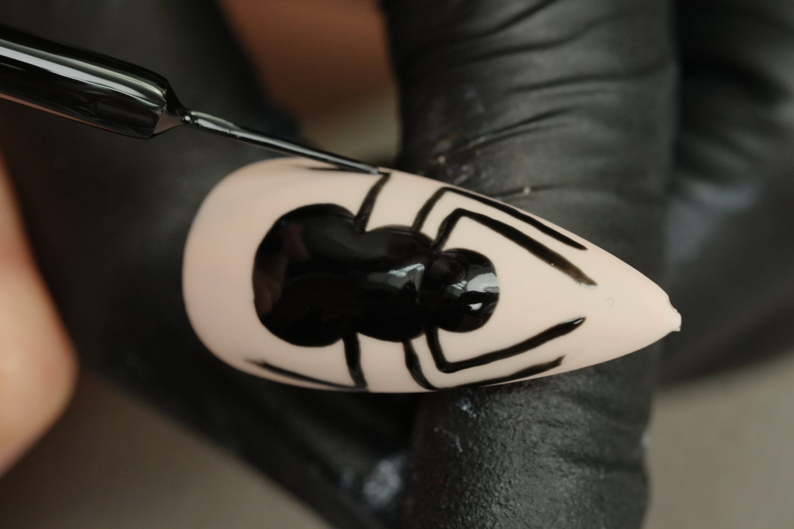 Halloween Nail Art: A close-up of a hand with a black glove carefully painting a black spider on a beige artificial nail. The person is using a fine-tipped brush to outline the spider’s legs and body, highlighting the precise work required to create detailed spooky designs.