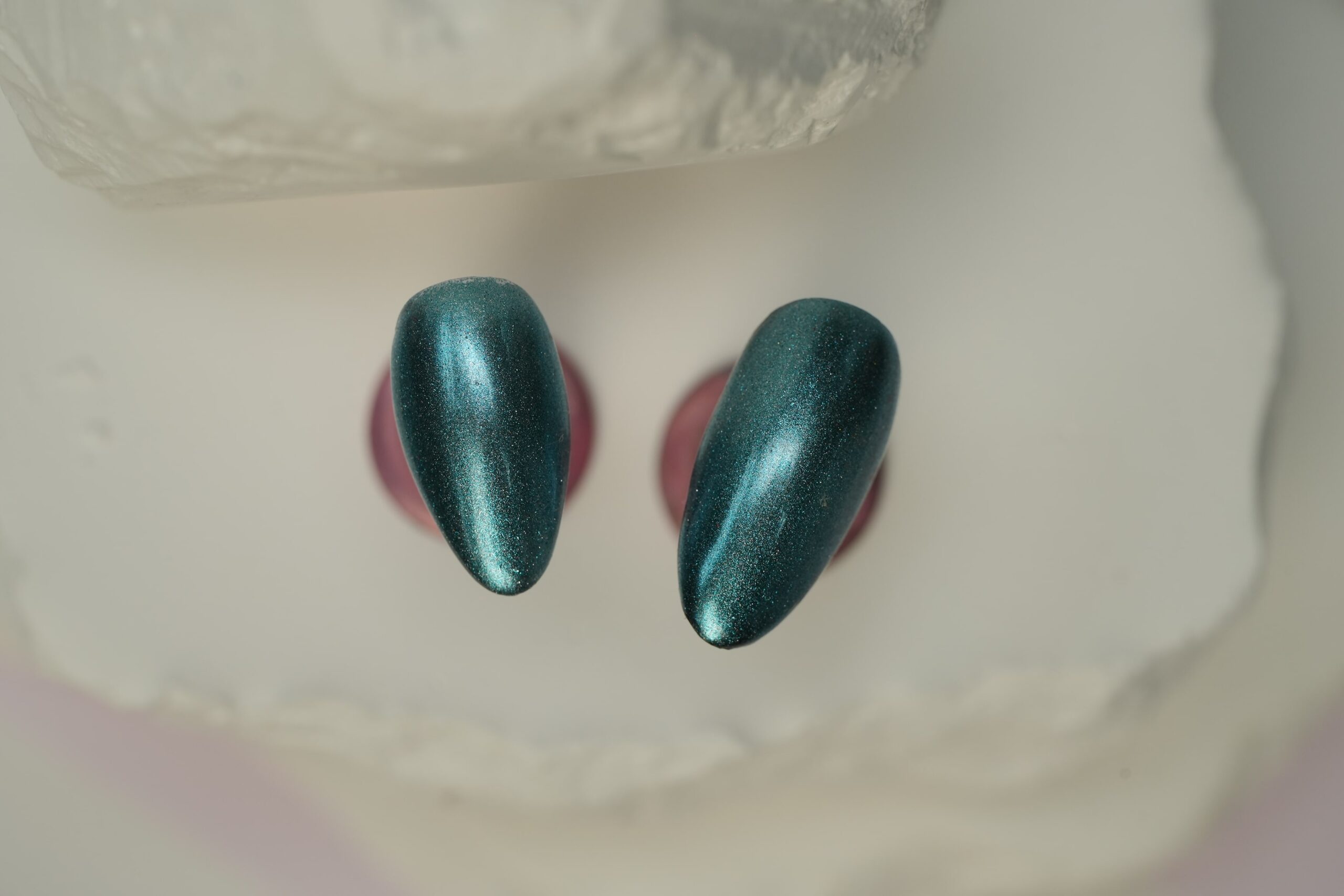 A pair of finished, almond-shaped nail tips with a deep teal chrome effect, showcasing a shiny, metallic finish.