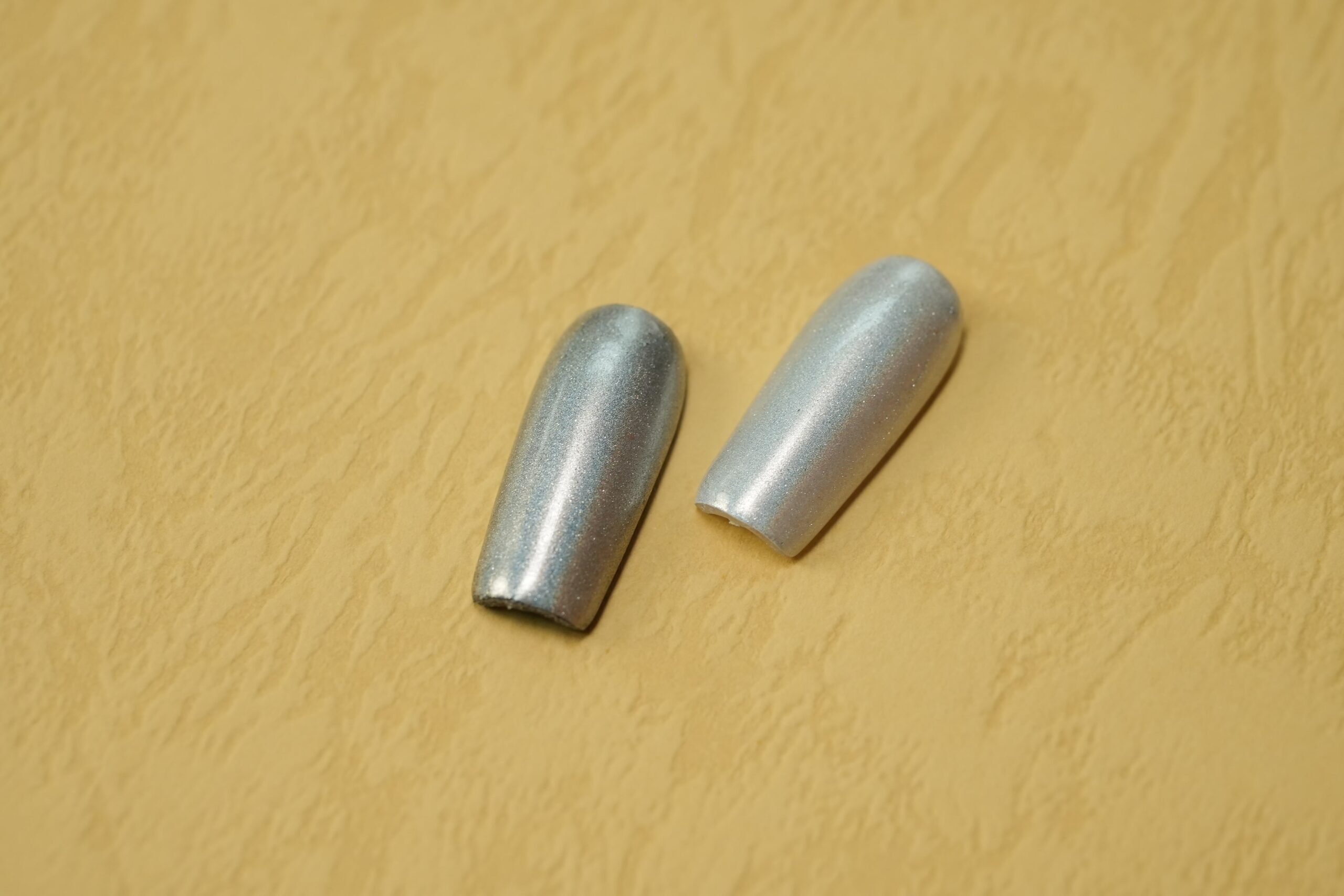 Two finished, coffin-shaped nail tips with a shimmering silver chrome effect, demonstrating a smooth, reflective surface.