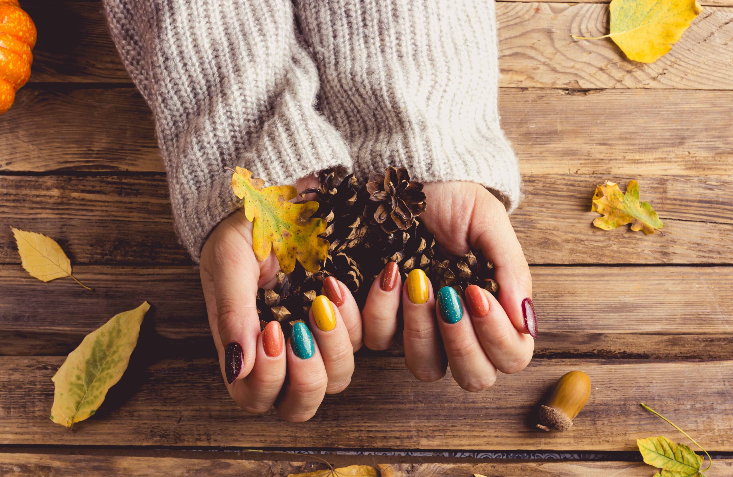 A person's hands, wearing a cozy grey sweater, are holding a cluster of pinecones on a rustic wooden table. Their nails are painted in various fall colors, including glossy dark red, glittery teal, orange, and yellow, highlighting a mix of creative and cozy color combinations for fall nail art.