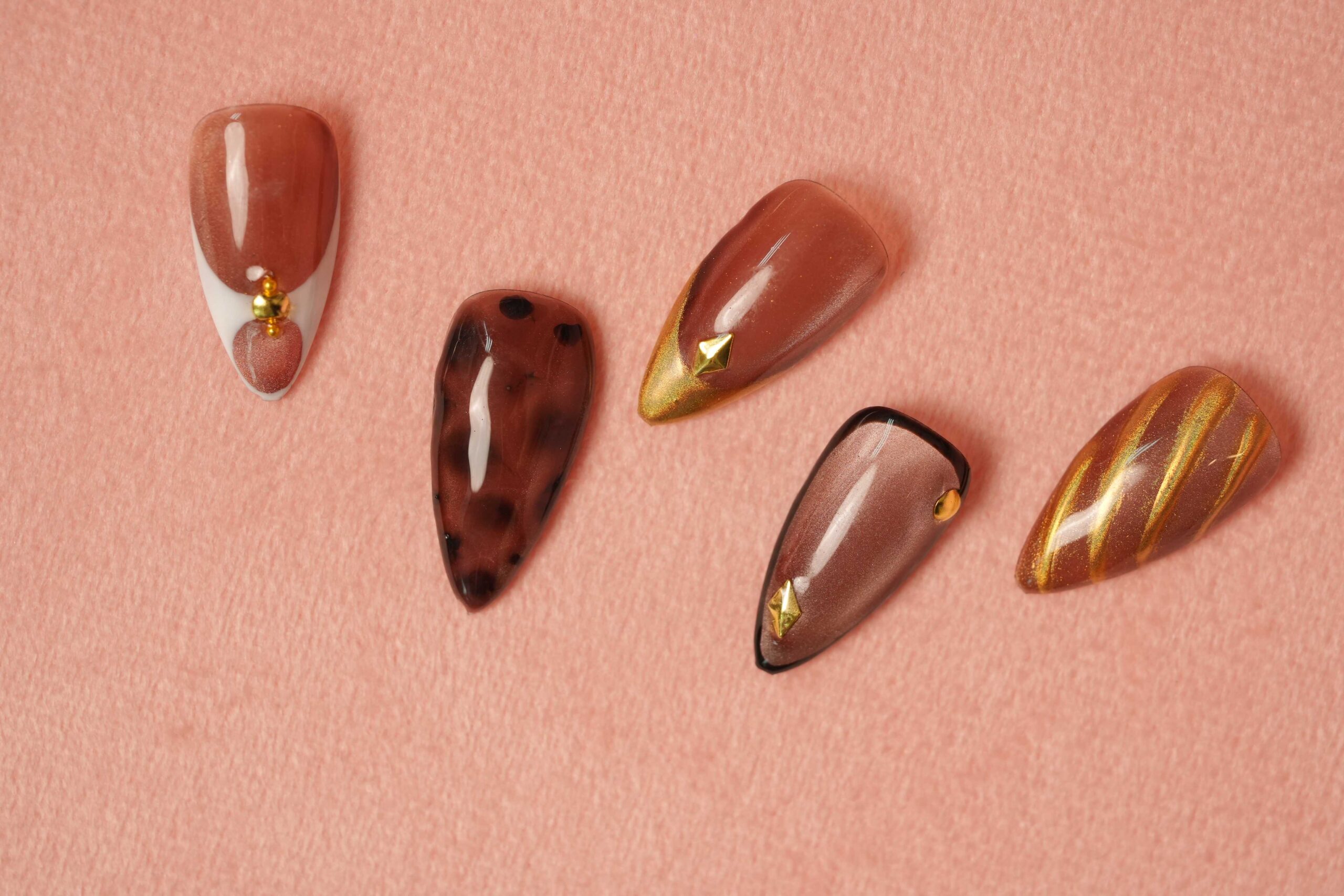 Five artificial nails with different designs for fall, arranged on a pink background. The nails feature creative ideas such as a maroon animal print, a gold French tip, a matte mauve with a metallic tip, and a brown marble effect, showcasing cozy and unique color combinations for fall nail art.