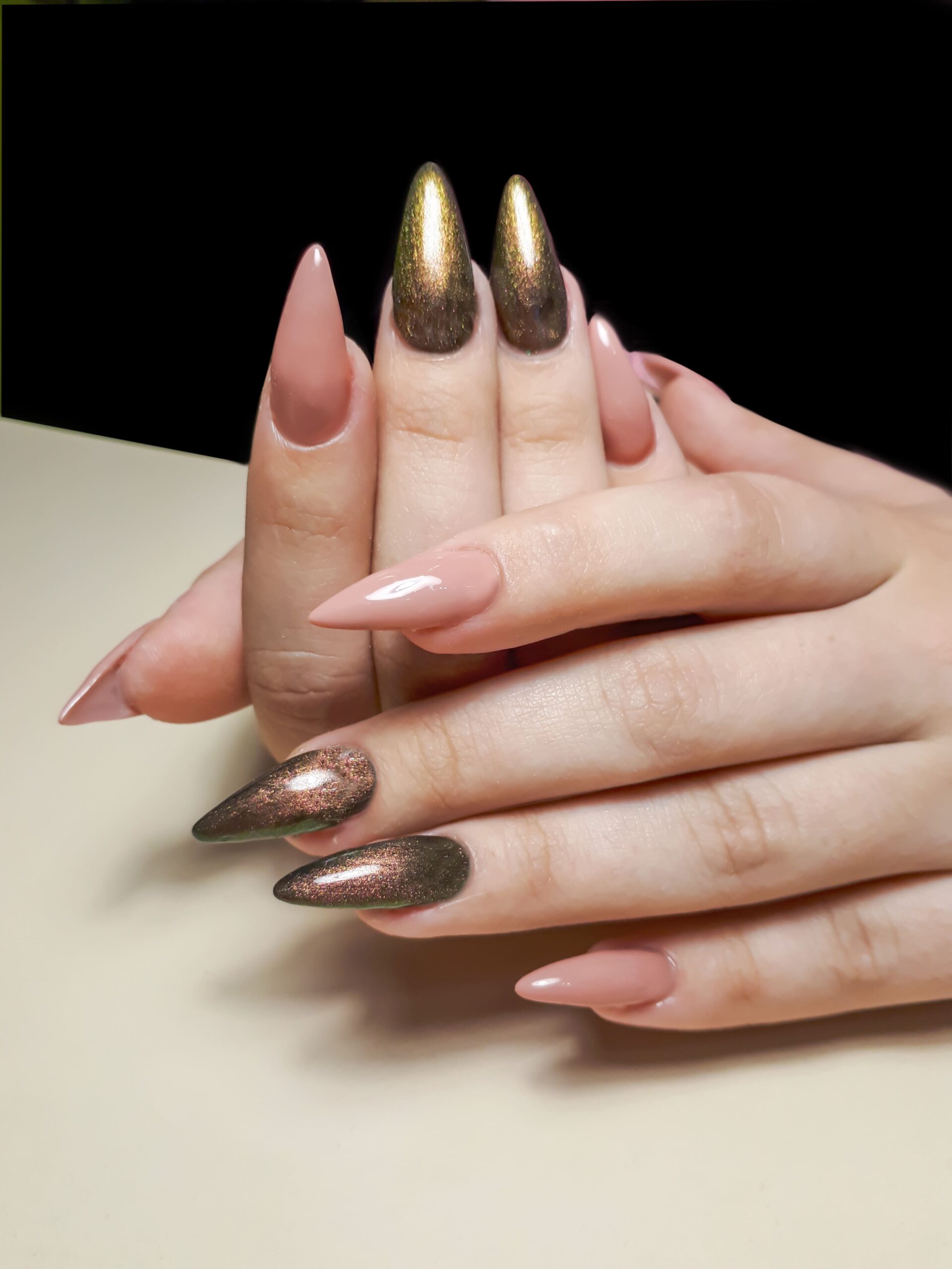 A close-up of a person's hands with long, almond-shaped nails. The nails are painted in a mix of glossy nude pink and a stunning golden-brown cat-eye polish, showing a versatile nail art look for both party nights and cozy events.