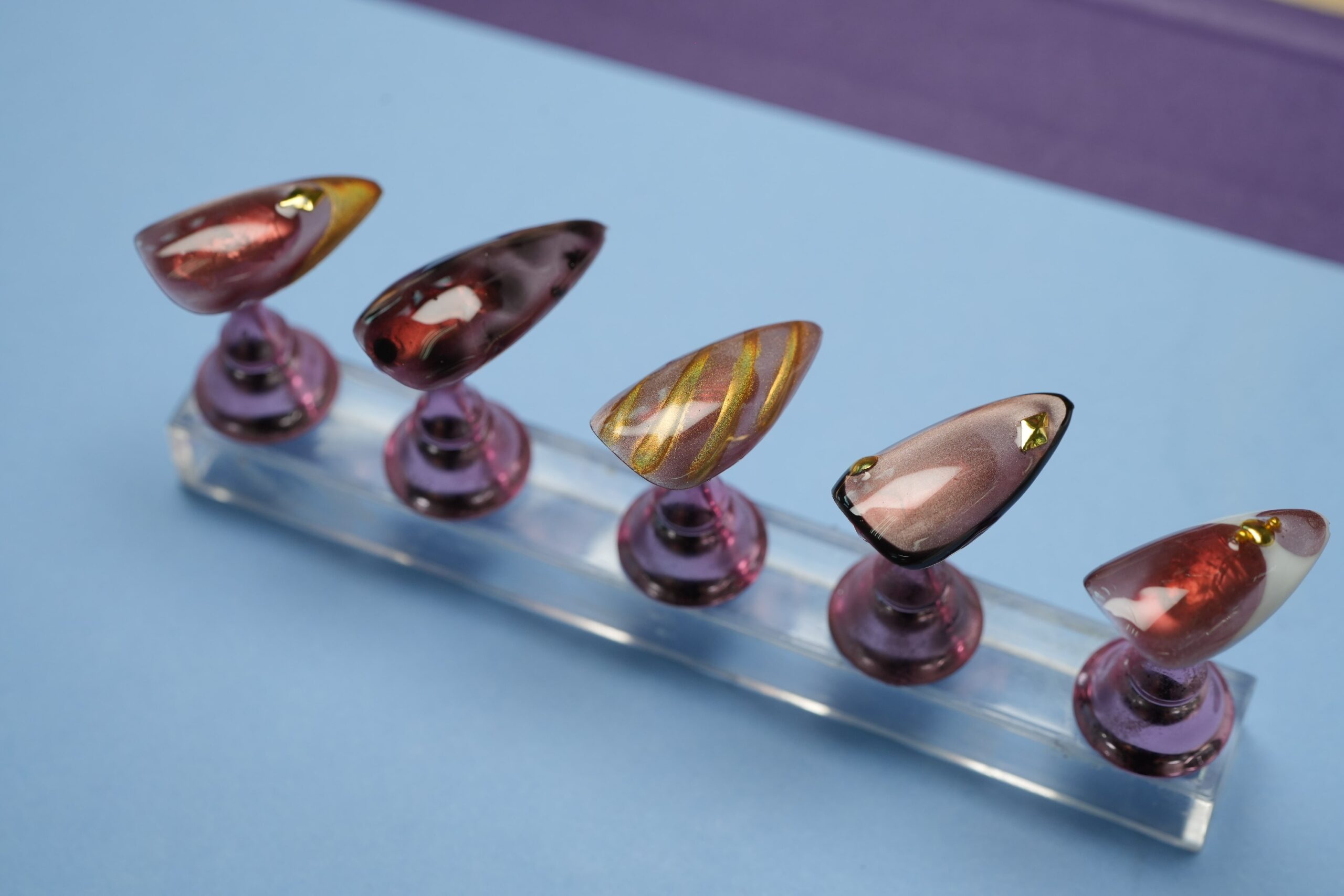 Five artificial nails displayed on a clear stand, showcasing creative nail art for fall events. The designs feature abstract swirls, metallic gold stripes, and gem accents, with colors ranging from deep brown and red to sheer nude, perfect for various fall occasions.