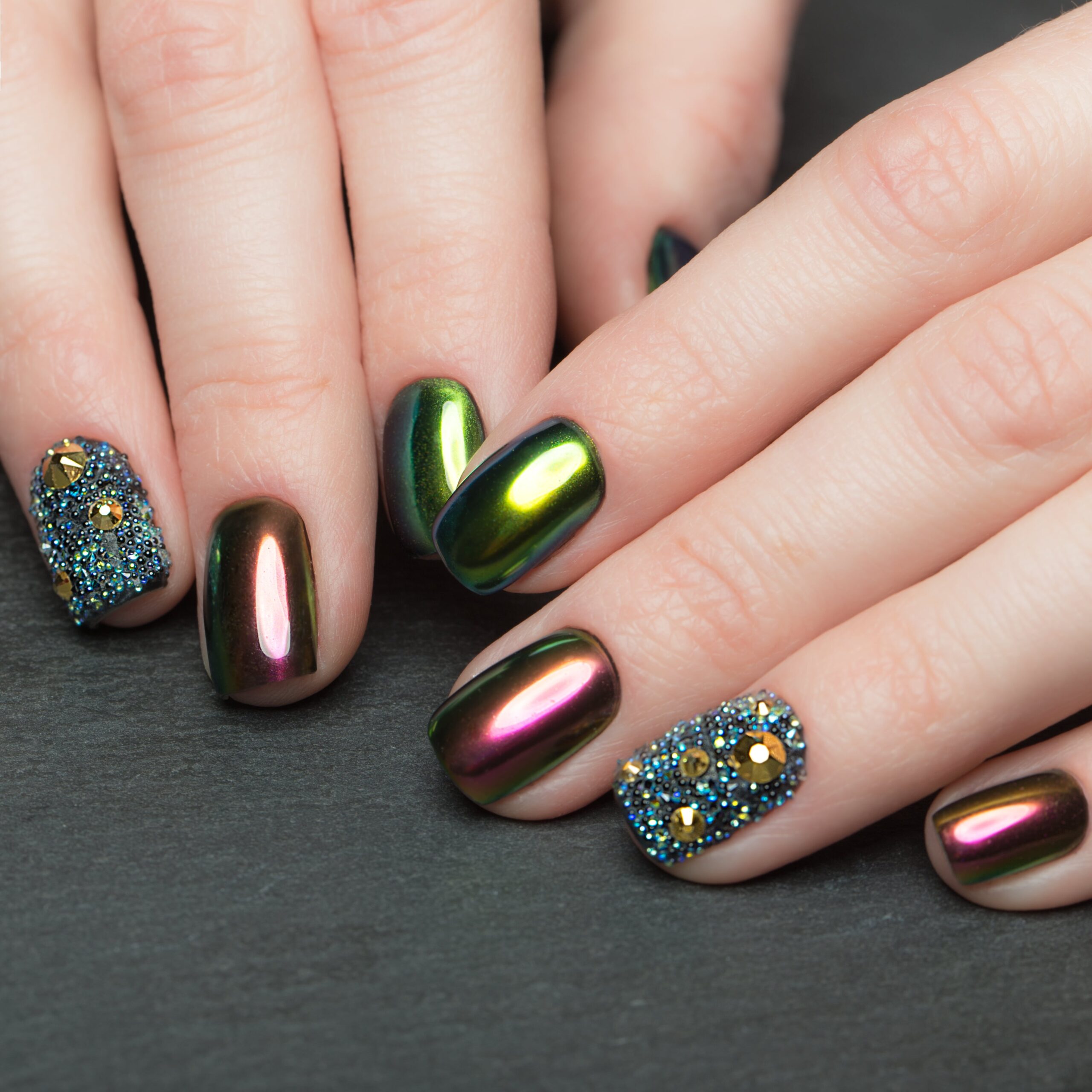  A close-up of hands with bold, square-shaped nails featuring a dramatic fall party look. The nails are painted in a vibrant green and purple iridescent chrome finish, with two nails fully covered in sparkling blue and gold rhinestones, ideal for a glamorous fall event.