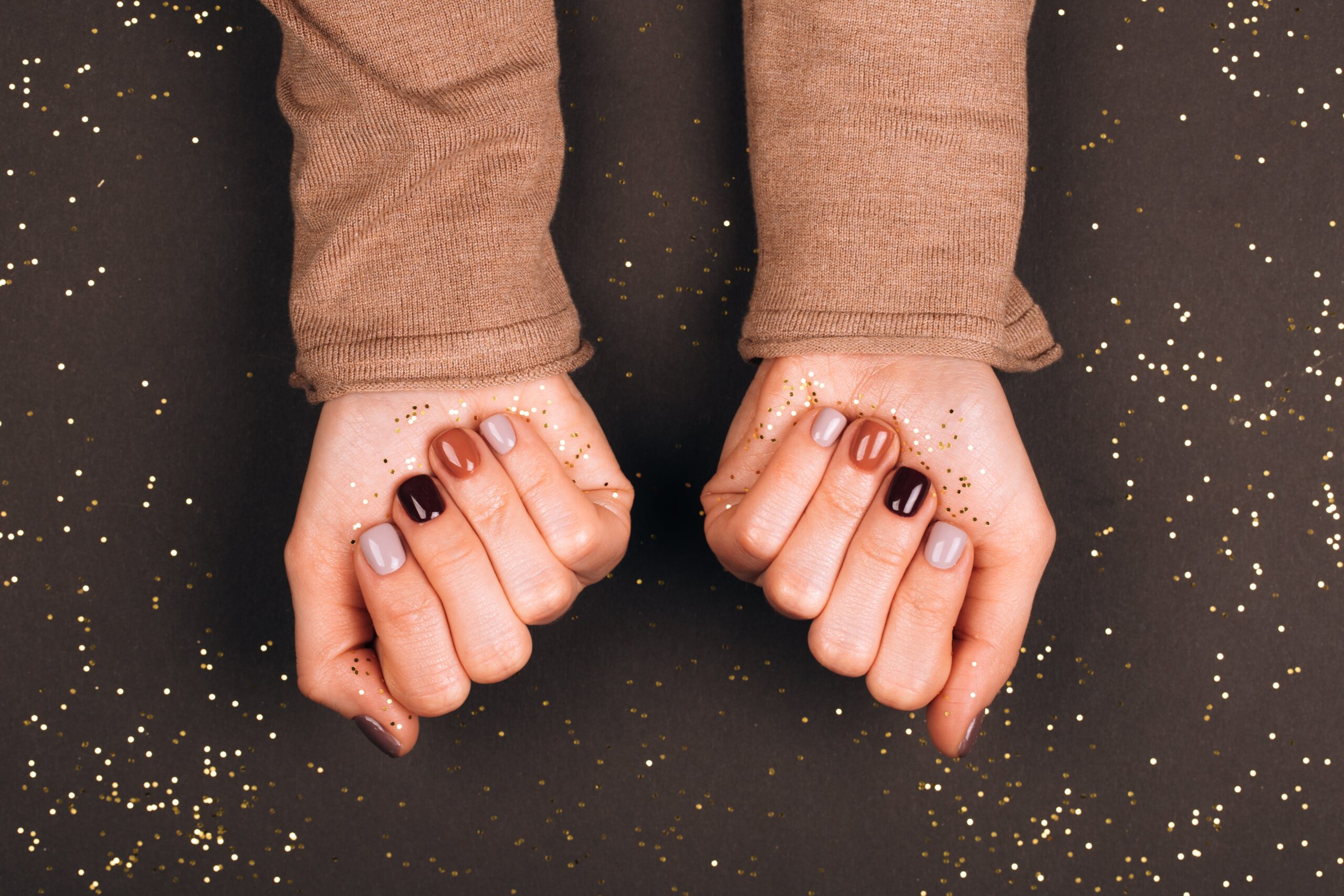 A person's hands, wearing a cozy brown sweater, are held over a dark background with scattered golden glitter. The nails are painted in a sophisticated fall palette of deep burgundy, earthy brown, and pale taupe, showcasing a perfect manicure for a cozy night in.