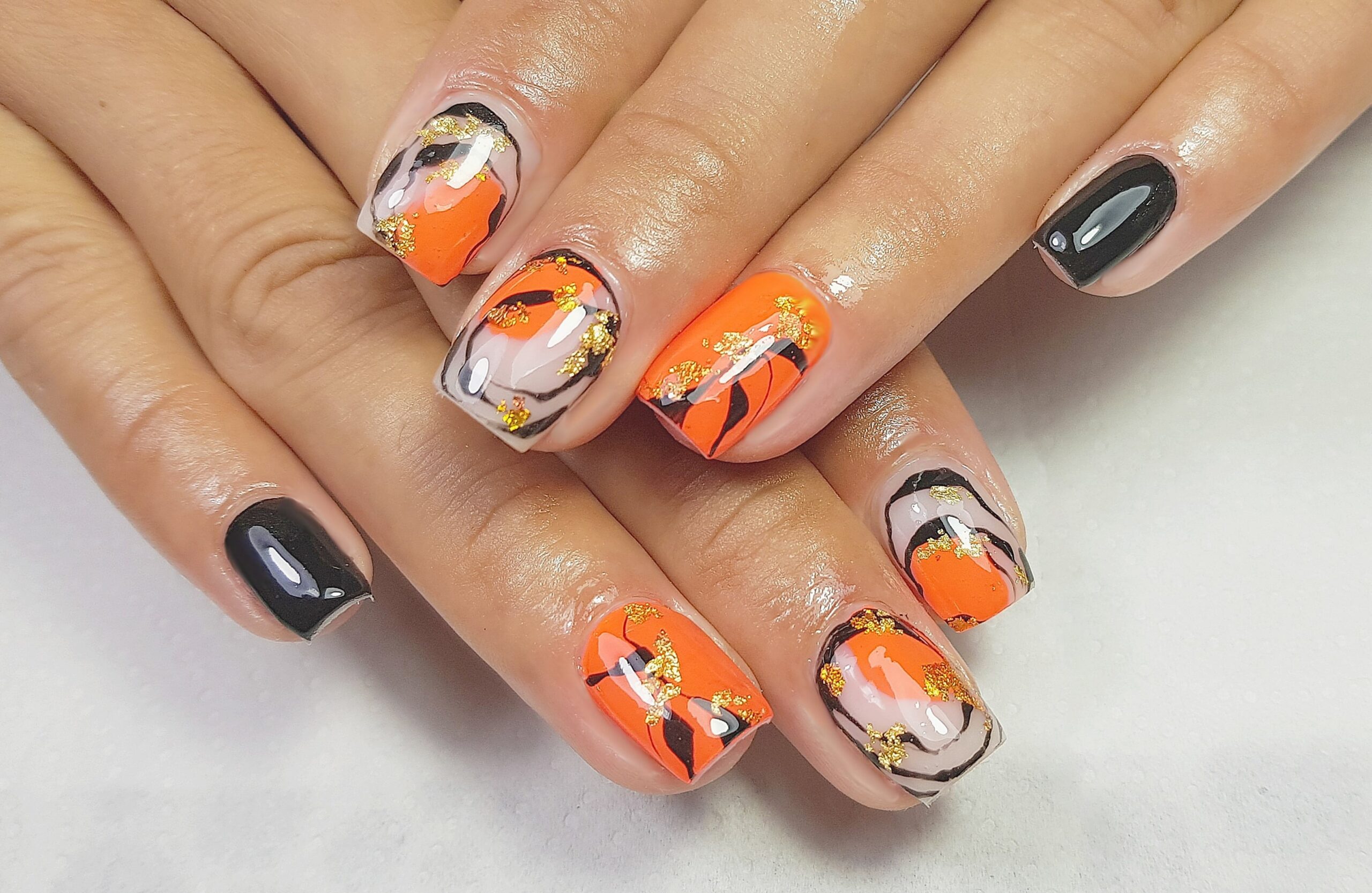 A close-up of hands with short, square-shaped nails featuring a dynamic fall nail art design. The nails are painted in a high-contrast pattern of bright orange, black, and white swirls with gold foil flakes, offering a creative and bold look for a fall party.