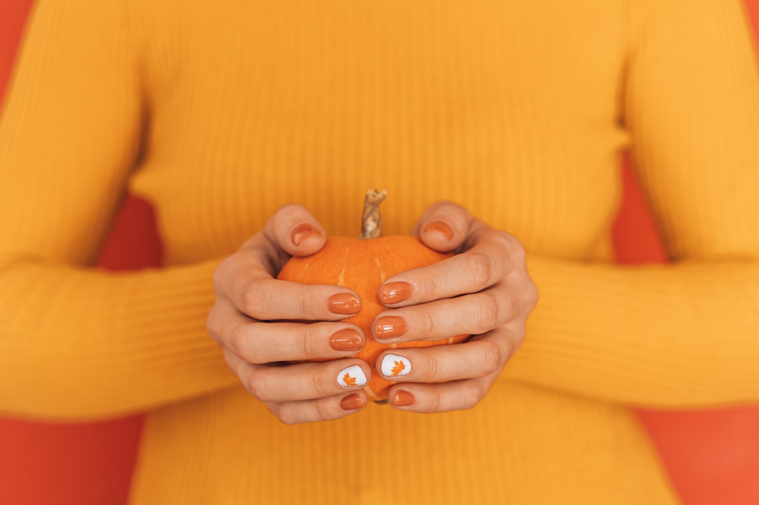A person wearing a yellow sweater holds a small orange pumpkin. Their nails are painted in a glossy orange, with a hand-painted leaf on one accent nail, showcasing a simple yet chic design perfect for a casual fall event or cozy activity.