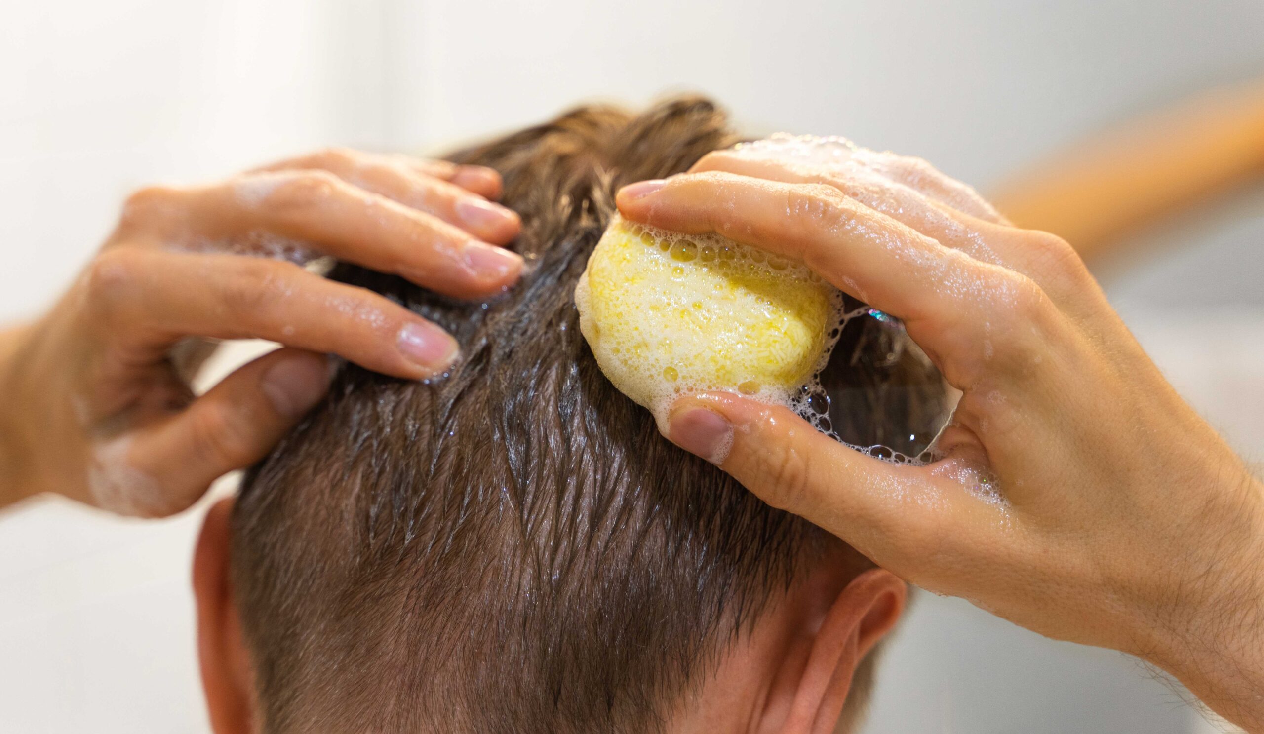 A person's hands lathering a yellow shampoo bar into wet hair, suggesting different product formats for treating hair loss.
