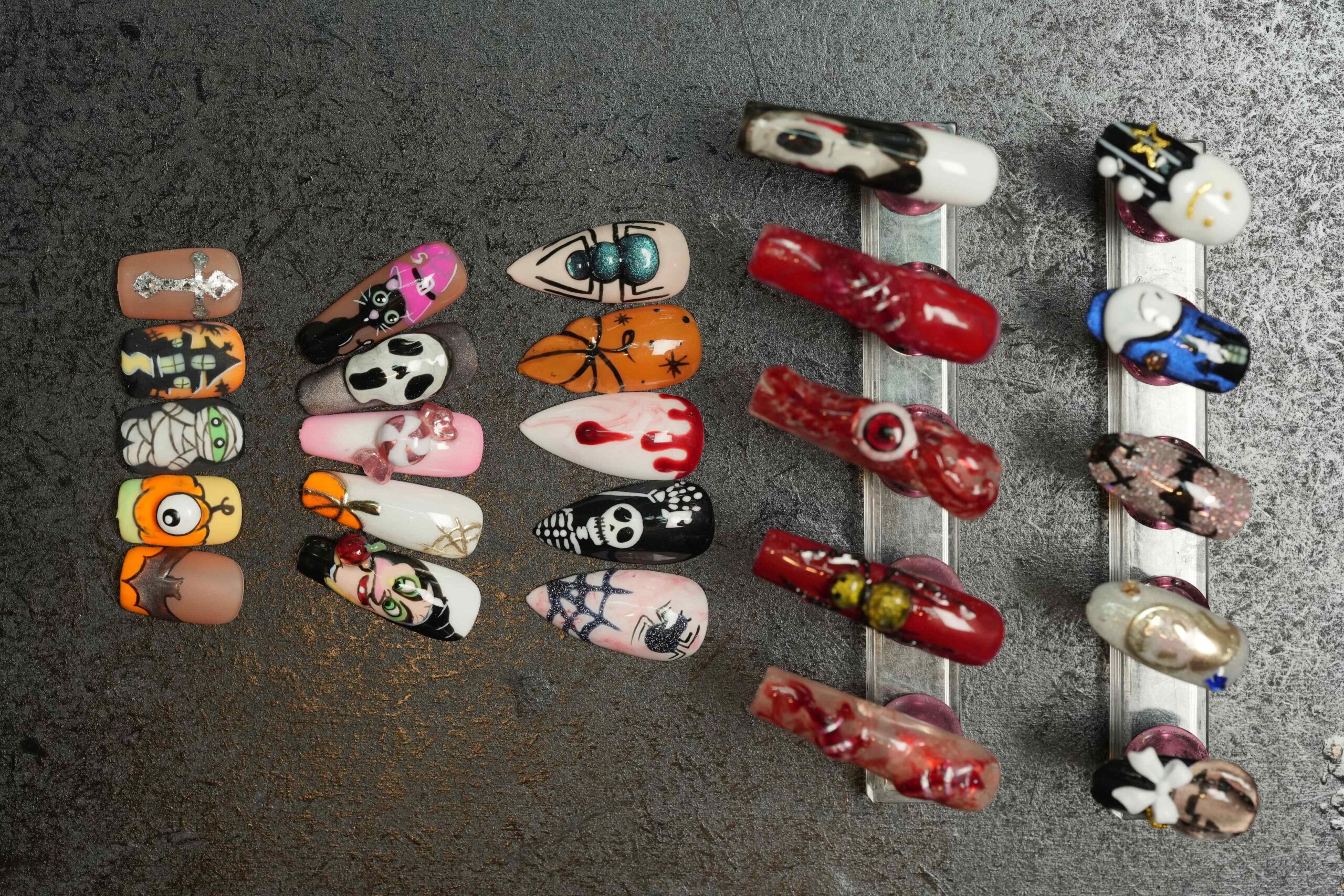 Halloween Nail Art: A comprehensive collection of artificial nails arranged on a dark metallic surface. The designs include mummies, pumpkins, bats, ghosts, skeletons, and dripping blood, illustrating a wide range of creative ideas and spooky color combinations for Halloween nail art.