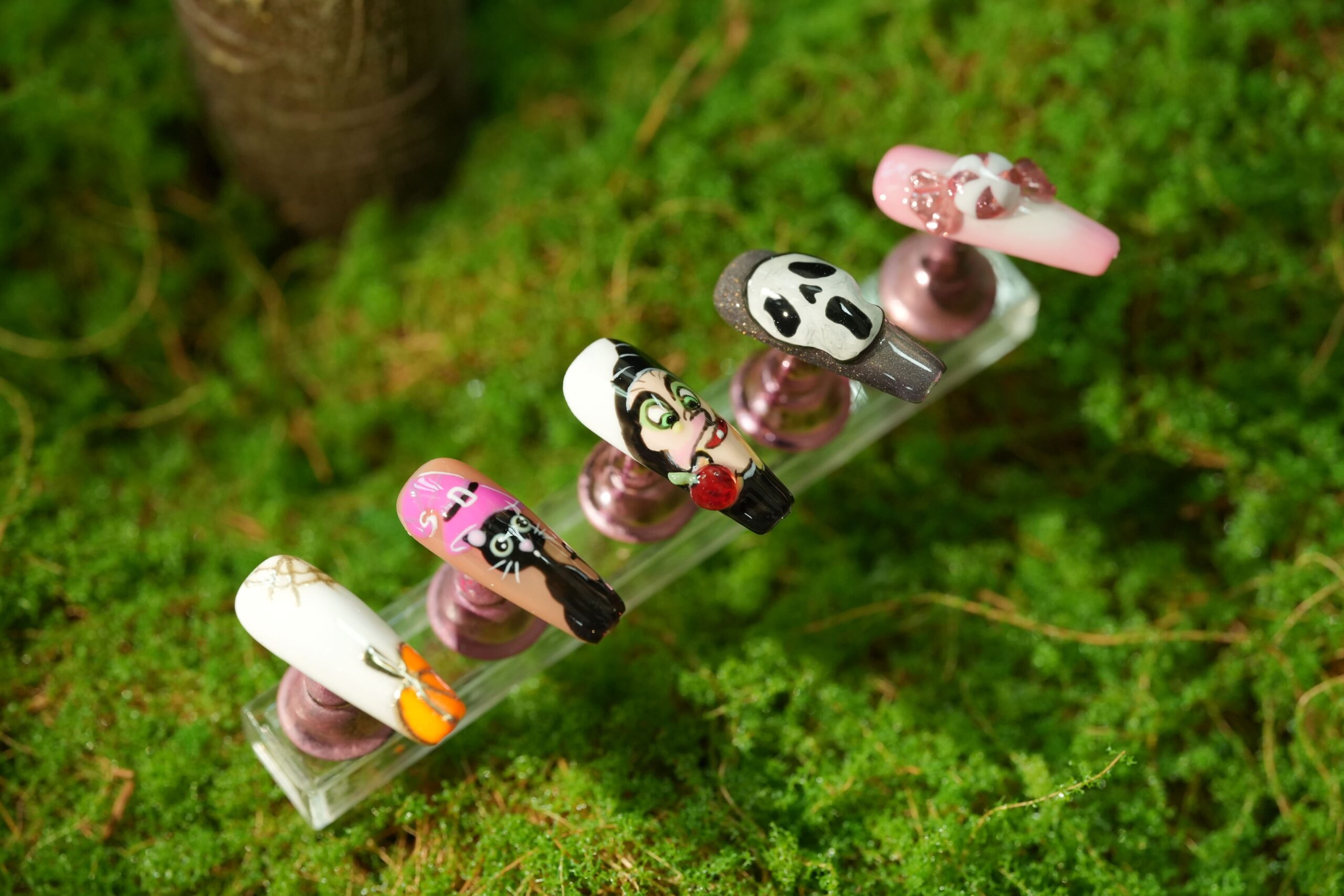 A set of artificial nails displayed on a bed of green moss. The designs feature cartoon-like characters including a ghost, a black cat, a vampire character, and a skull, showcasing creative ideas and spooky color combinations inspired by classic Halloween figures.