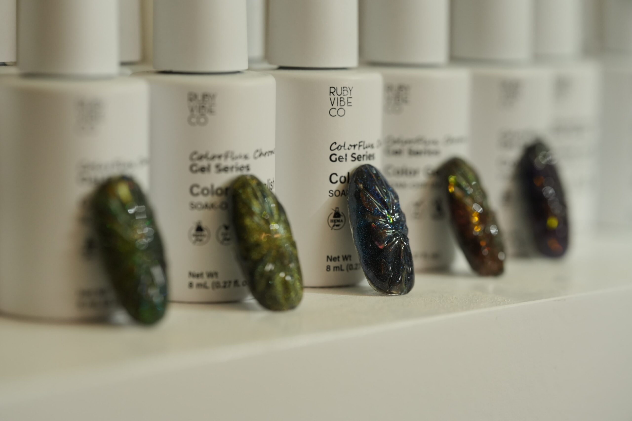 Halloween Nail Art: A row of unique, insect-inspired artificial nails placed in front of a line of Ruby Vibe Co. gel polish bottles. The nails have a special iridescent finish resembling beetle wings, highlighting spooky and creative color combinations and ideas for Halloween nail art.