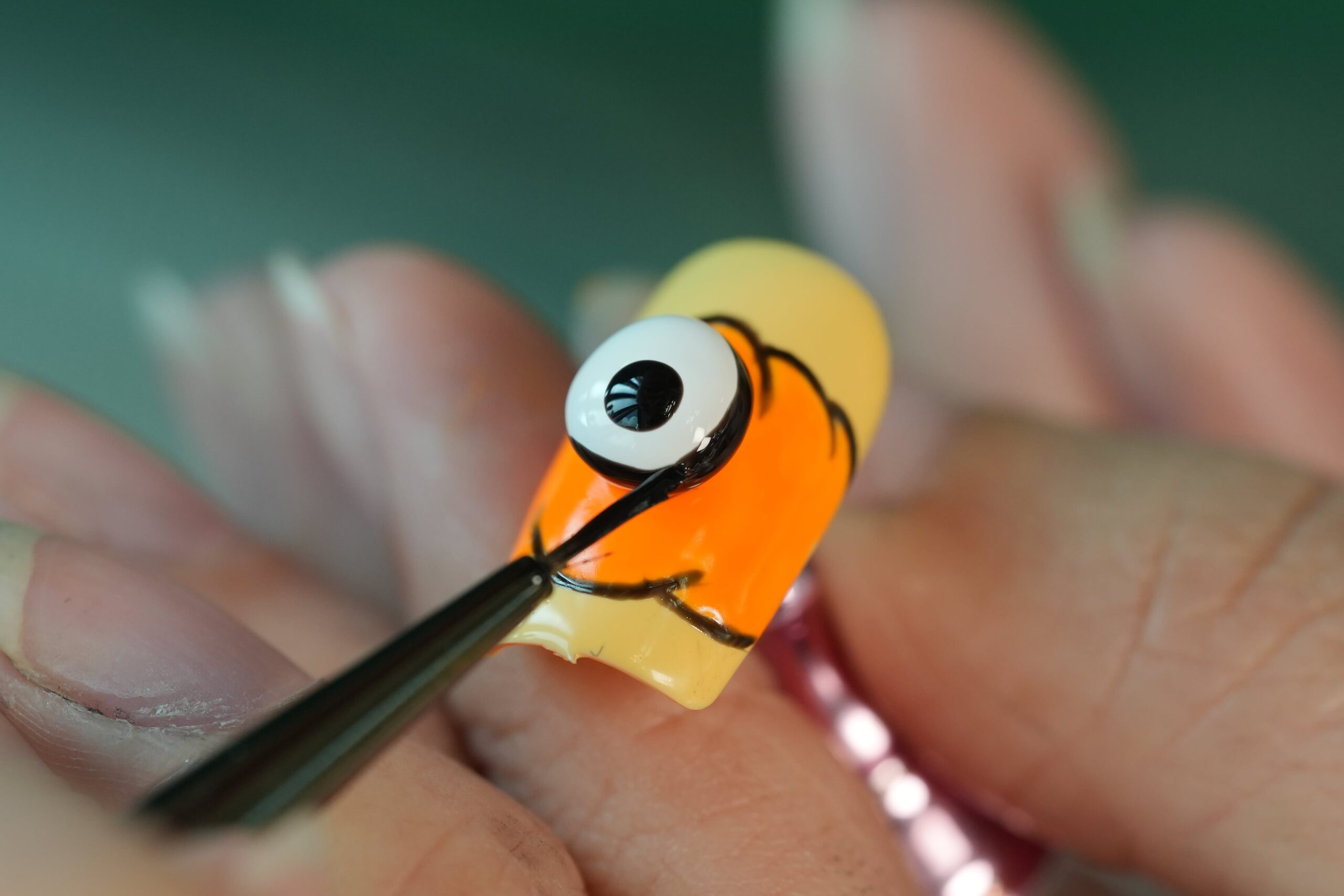Halloween Nail Art: A close-up of a person using a fine brush to create a creepy eyeball design on an artificial nail. The nail features a bold orange and yellow base with a prominent 3D eyeball, highlighting a creative and spooky nail art idea for Halloween.