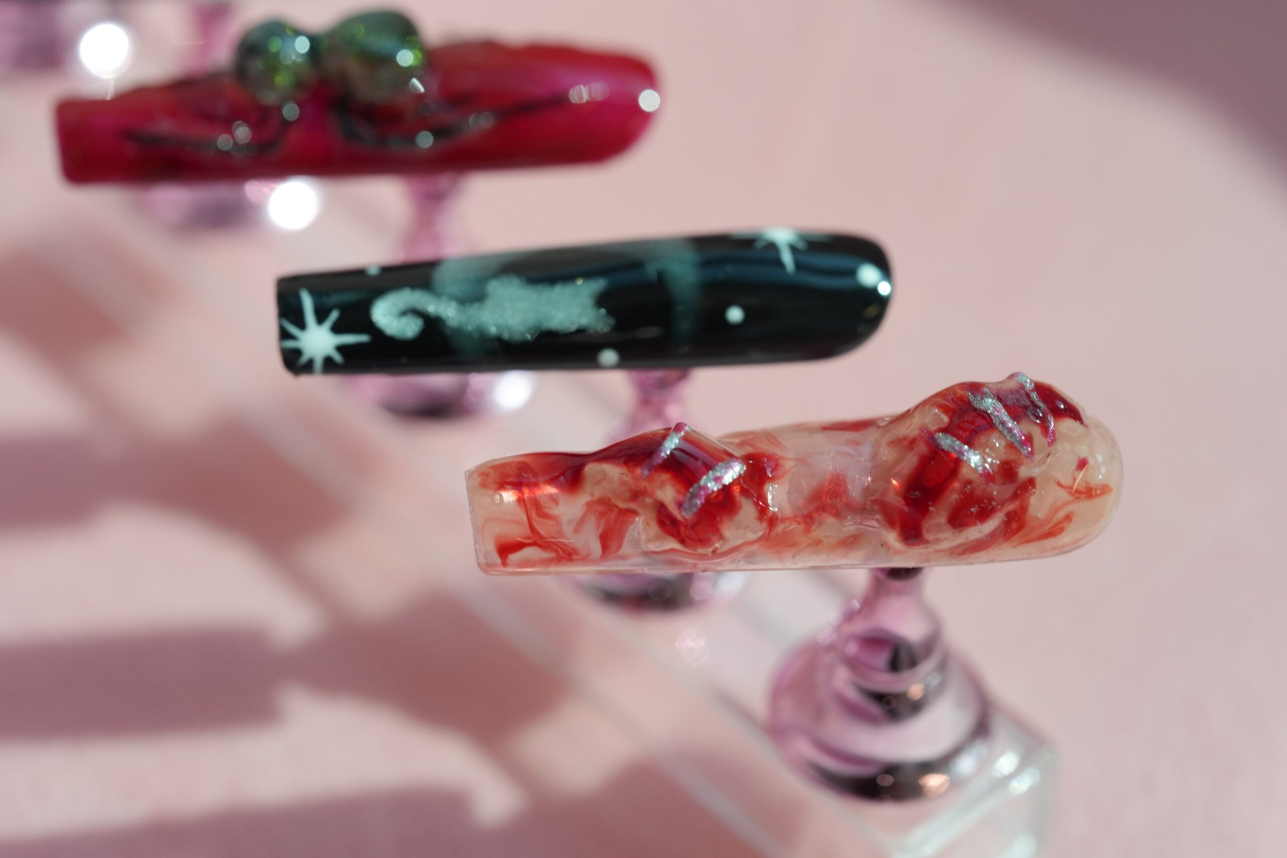 A close-up of three artificial nails with horror-themed designs. One nail has a blood-spatter pattern with silver glitter accents, another features a cat and stars on a night sky background, and a third is a deep red with decorative beads. These showcase creative and spooky ideas for Halloween nail art.