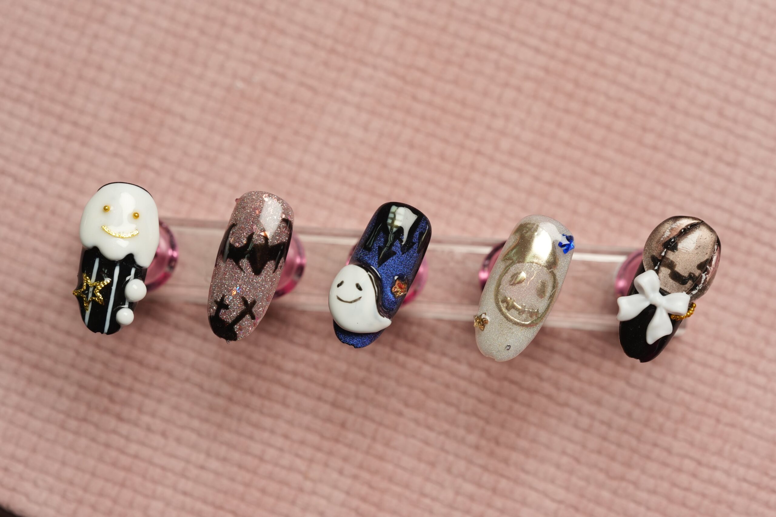 A set of five ghostly artificial nails with spooky designs. The designs include cute ghosts, ghouls, and a smiling face, adorned with small details like bows and stars, showcasing creative and fun Halloween nail ideas with unique color combinations.