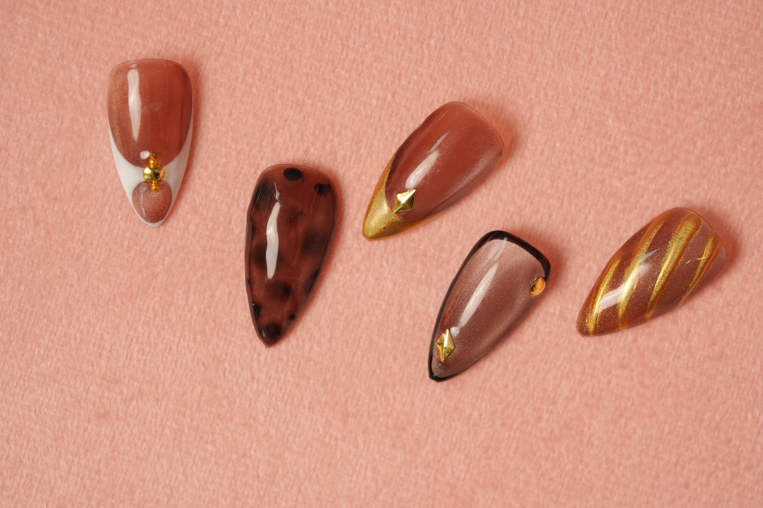 A set of five artificial nails in warm, earthy tones of brown and light pink. The designs include leopard print, gold shimmer, and pyramid studs, offering a sophisticated and cozy take on Halloween nail art with unique color combinations.