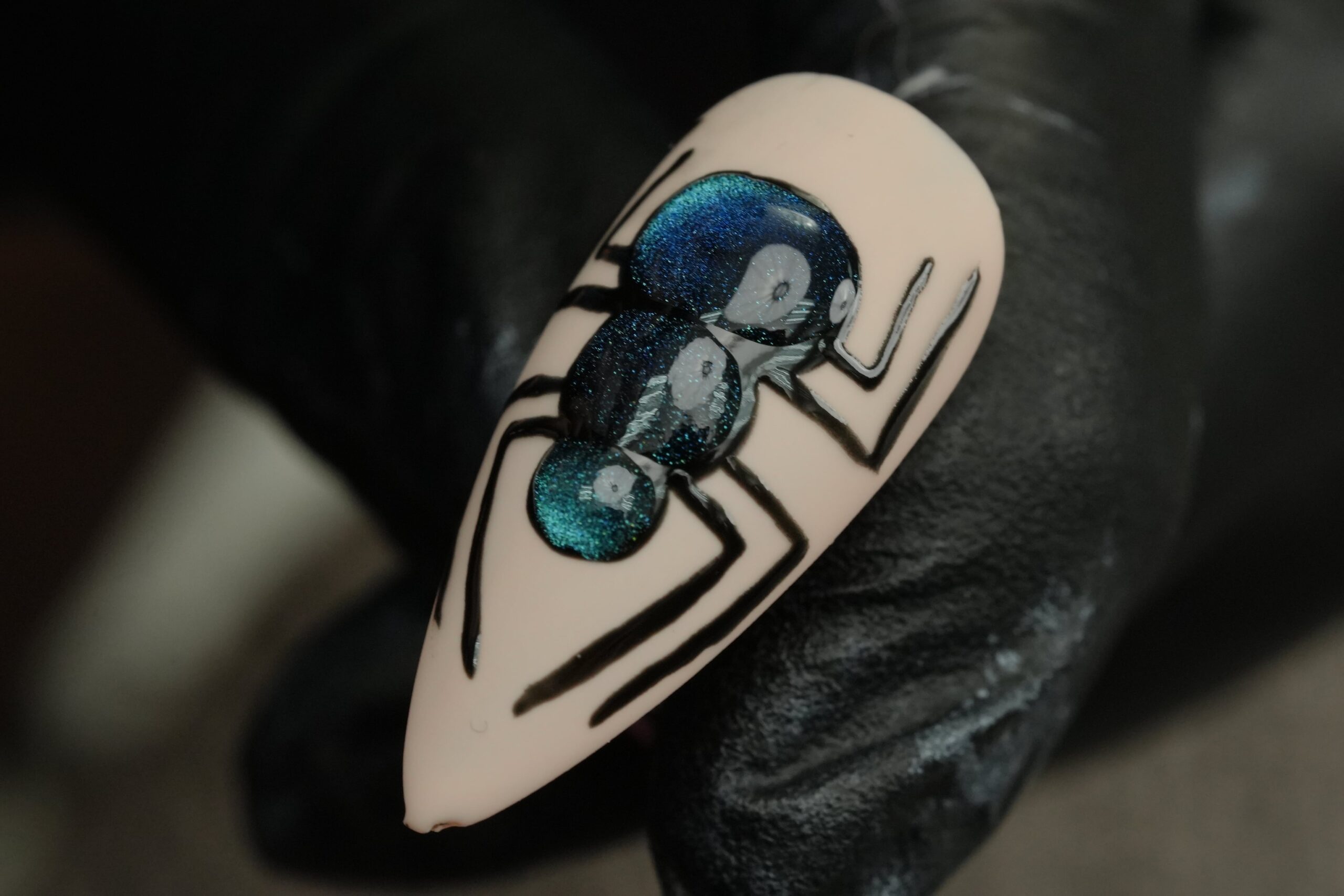 A close-up of a person wearing a black glove holding a beige artificial nail. The nail features a detailed black spider with a stunning iridescent blue and green finish on its body, showing a creative way to match a spooky Halloween costume.