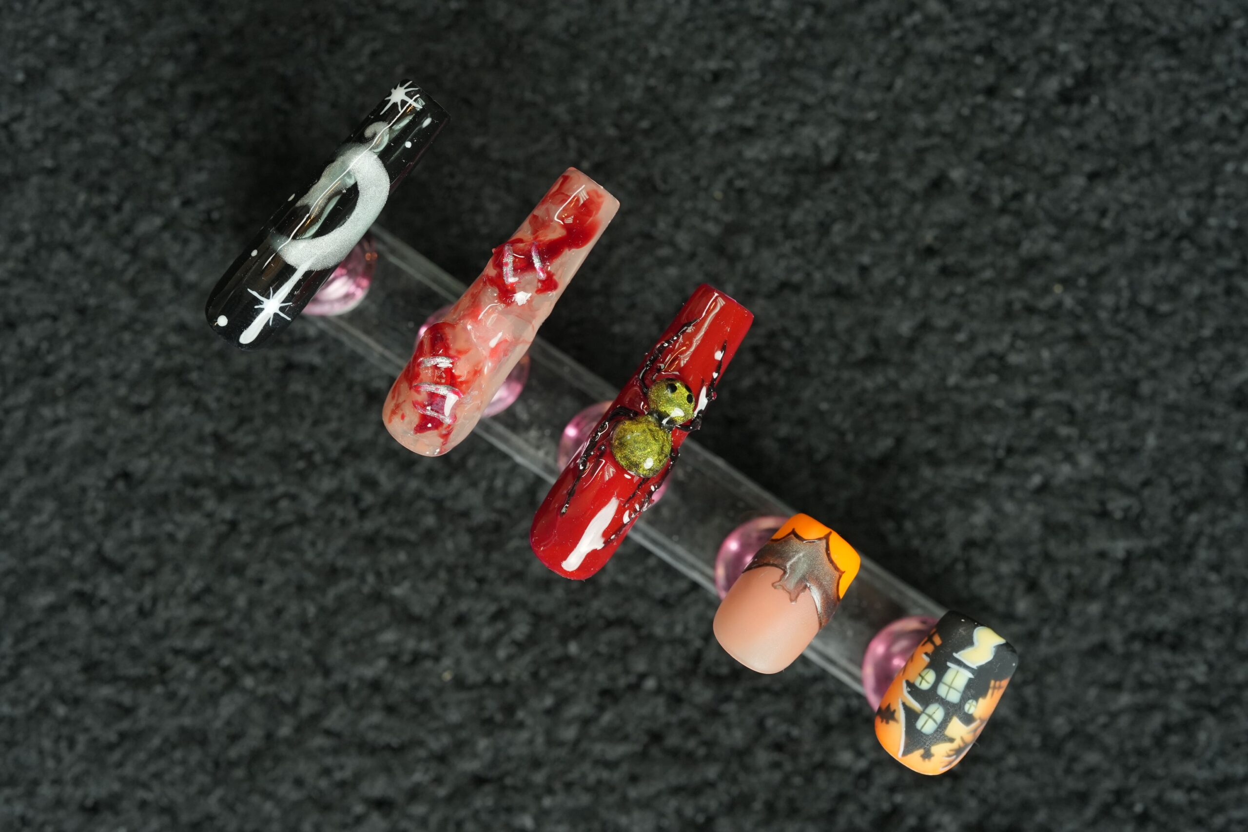Four artificial nails displayed on a clear stand with a dark, textured background. The nails feature different designs, including a black nail with a white moon, a bloody nail, a red nail with a creepy eye, and a nail with a haunted house scene, showcasing various options to perfectly match your Halloween costume.