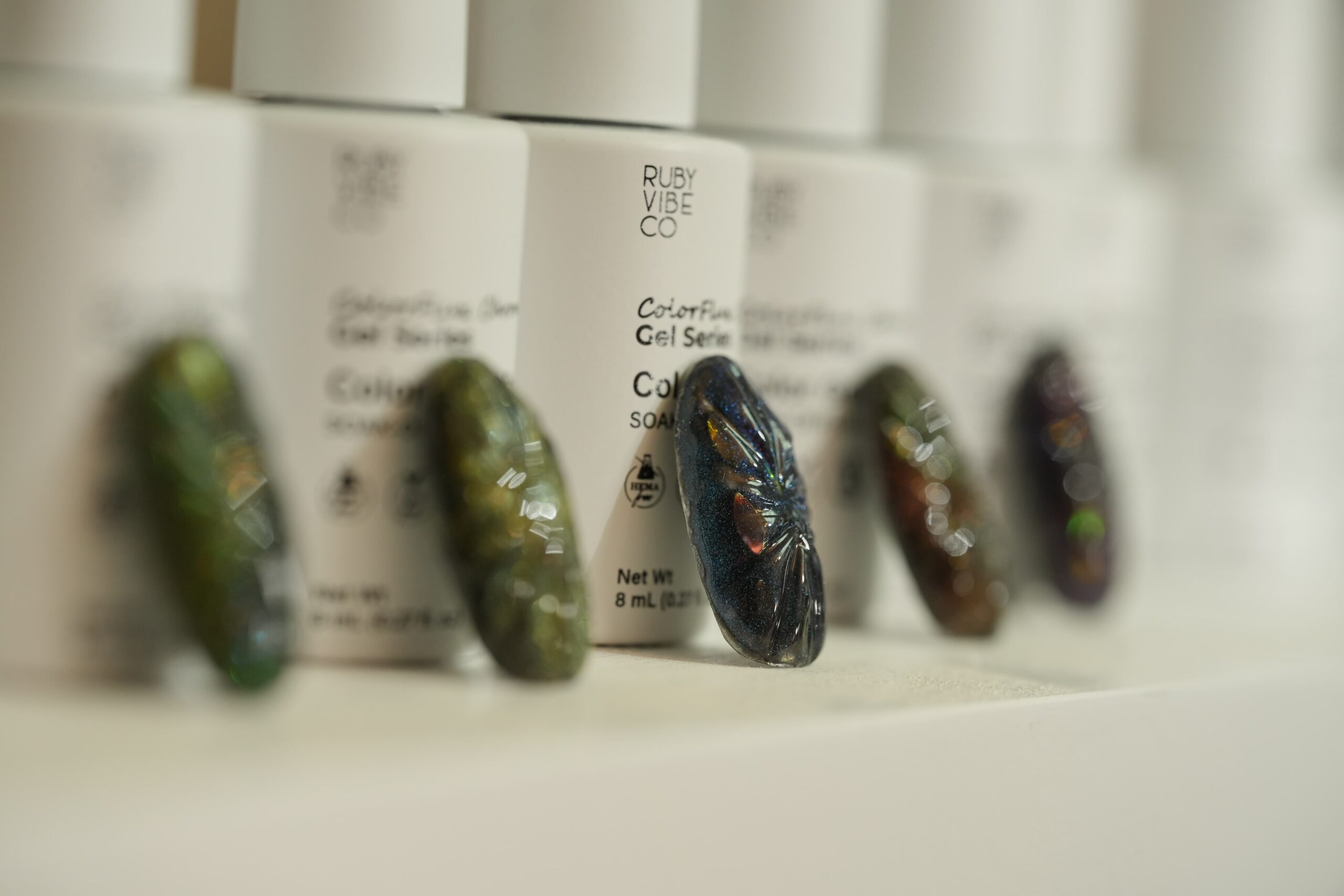  Halloween Nail Art: A row of iridescent, beetle-inspired artificial nails placed in front of several Ruby Vibe Co. gel polish bottles. The nails feature a rich, dark blue and black color combination with a shimmering finish, illustrating a unique and spooky way to match your costume.