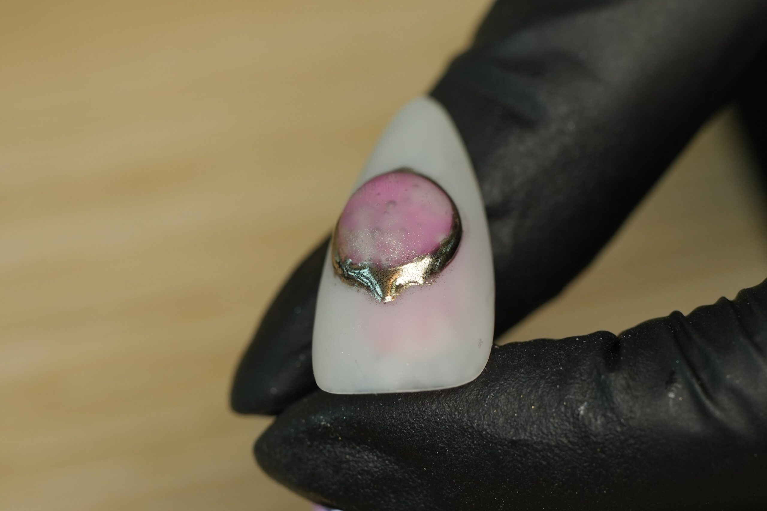 A close-up of a person wearing a black glove holding a pointed artificial nail. The nail features a pink base with a metallic, iridescent green and gold-colored foil detail on top, illustrating a stylish and creative accent to match a Halloween costume.