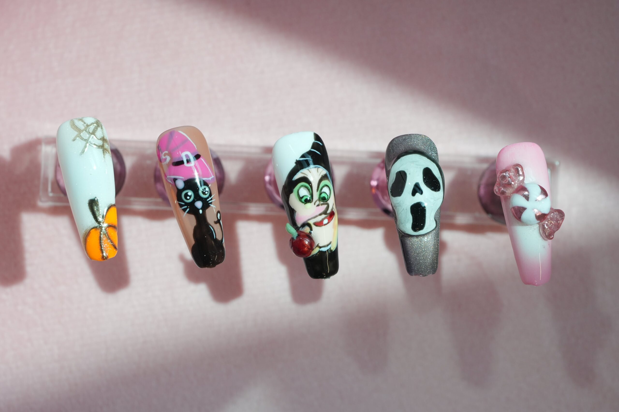A set of five artificial nails with various character-inspired designs, including a spooky pumpkin, a black cat with a witch hat, a vampire, a ghost face, and a pink design with a bow. The nails are displayed on a light pink background, showing a range of creative options to perfectly match your Halloween costume.