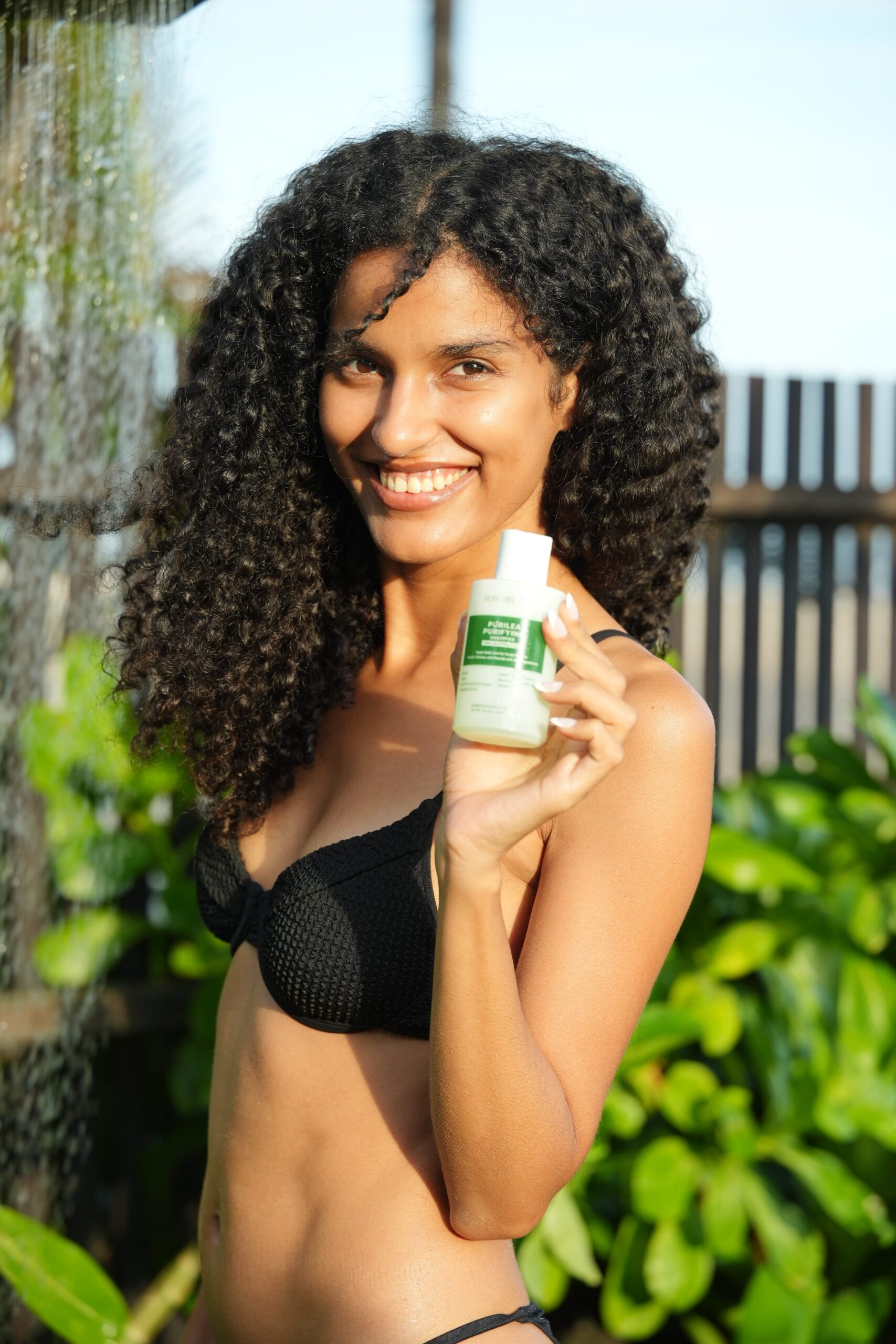 A smiling woman with wet, long, curly black hair holds a bottle of Ruby Vibe Co. Purilea Purifying Shampoo, suggesting she is happy with the product and its results for her hair.