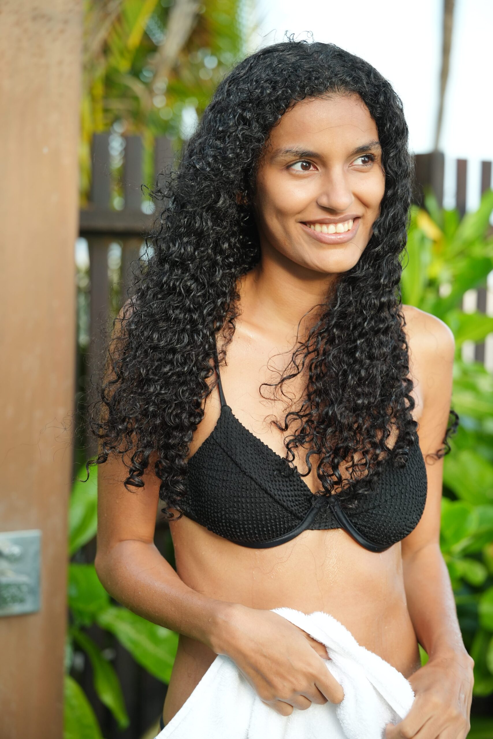 A woman with healthy, curly black hair stands outside smiling, holding a white towel. Her vibrant hair highlights the desired outcome of using a hair loss shampoo with effective ingredients.