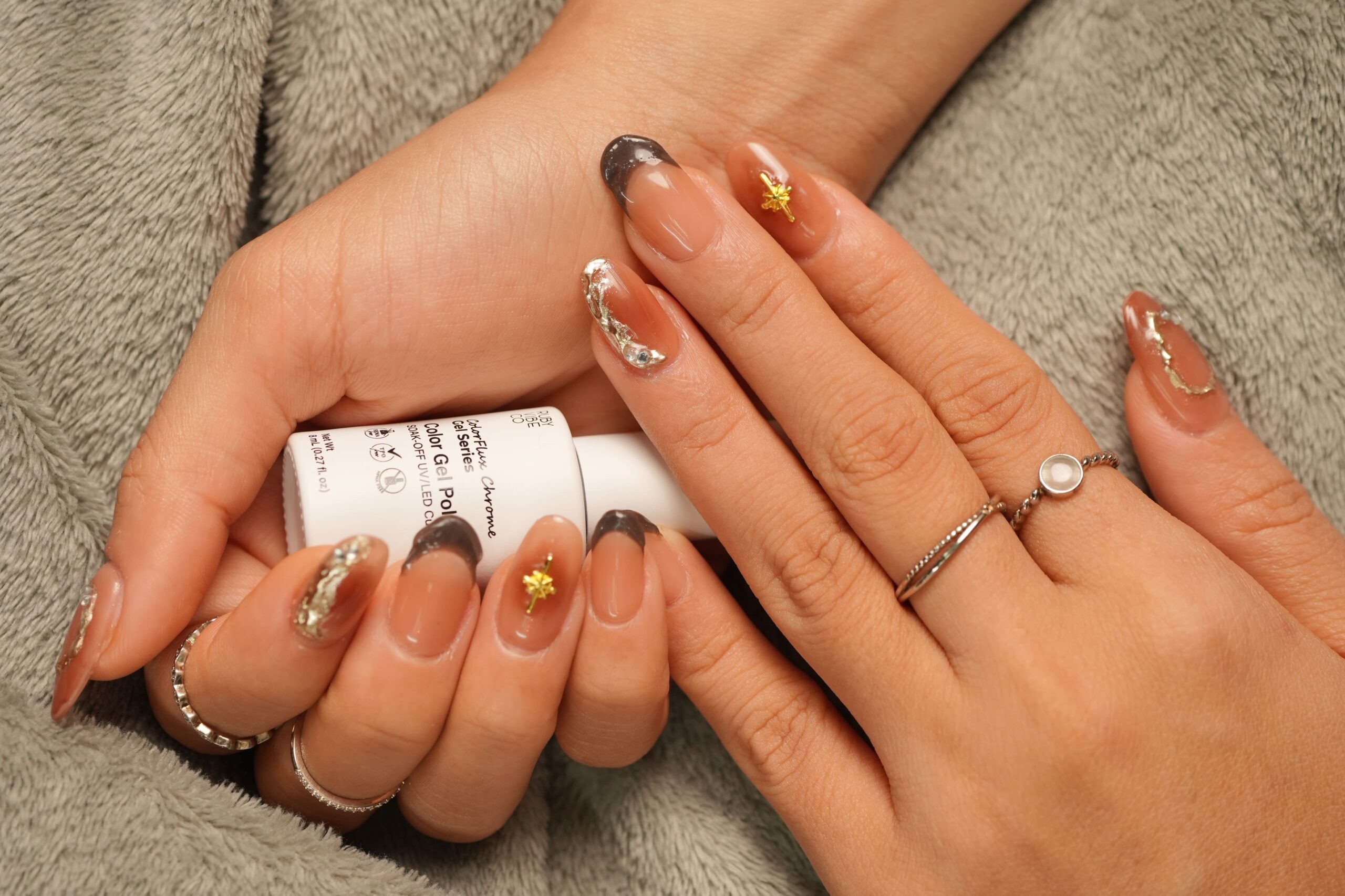A person holds a 'Ruby Vibe Co' gel polish bottle, showing off a long, elegant nail design with gold embellishments, a tip for a cozy autumn manicure.