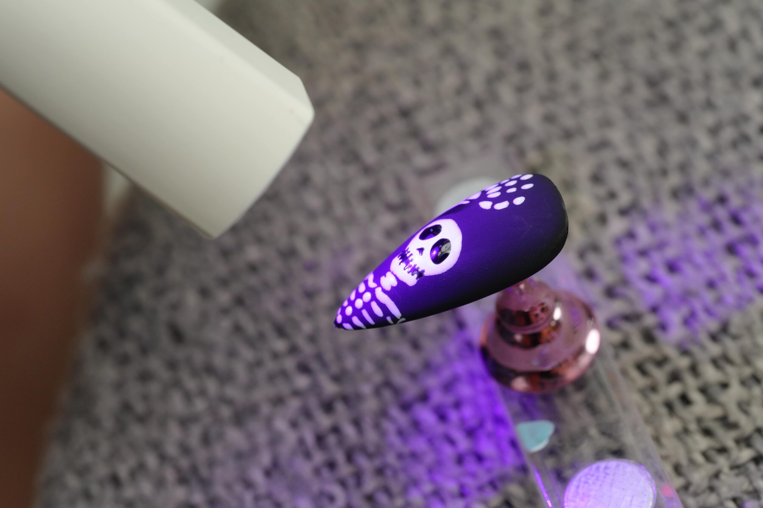 A black artificial nail with a white skull design is being cured under a UV lamp. The purple light accentuates the skull, demonstrating the gel curing process for durable, chip-free spooky Halloween nail art.