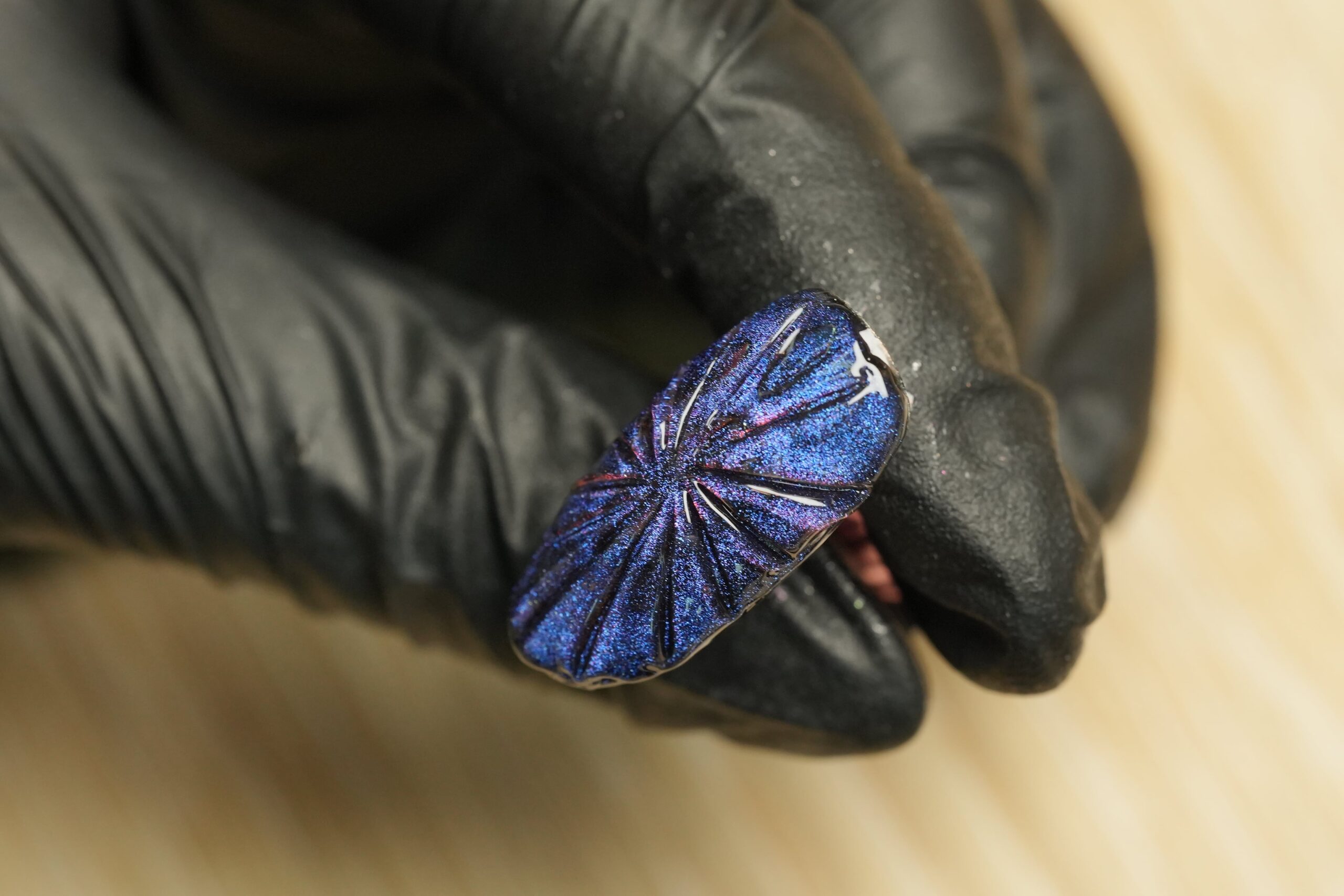  A gloved hand holds an artificial nail with an iridescent blue and purple gradient. The nail features textured, raised lines, illustrating a unique 3D nail art trick for creating a durable and visually striking spooky Halloween manicure.