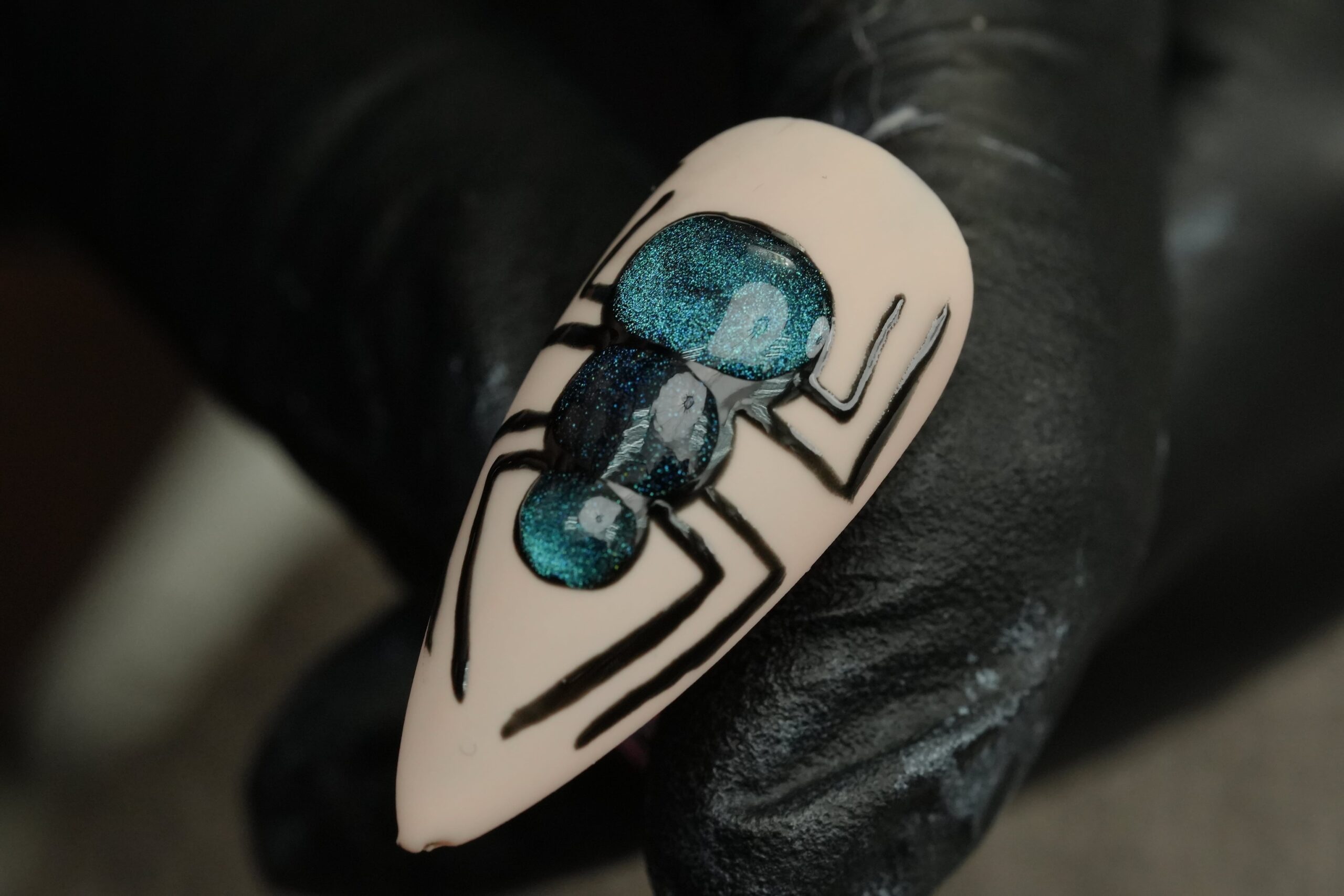 A close-up view of a detailed Halloween nail art design featuring a glittery, iridescent blue spider on a light nude nail base, held by a person wearing a black glove.