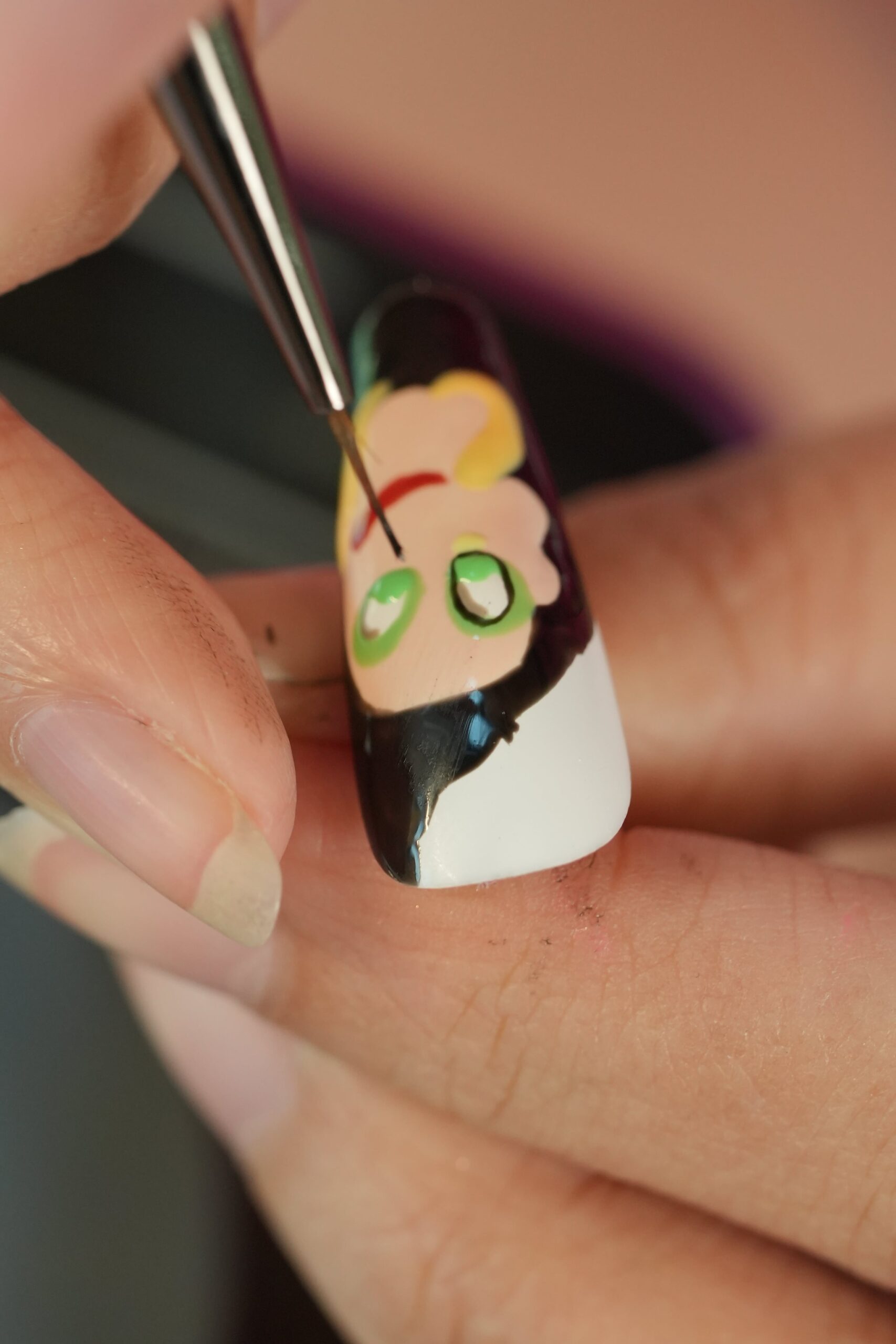 A close-up of a hand-painted Halloween nail design in progress, showing a manicurist using a fine detail brush to add features to a cartoon character with green eyes and a black jacket.