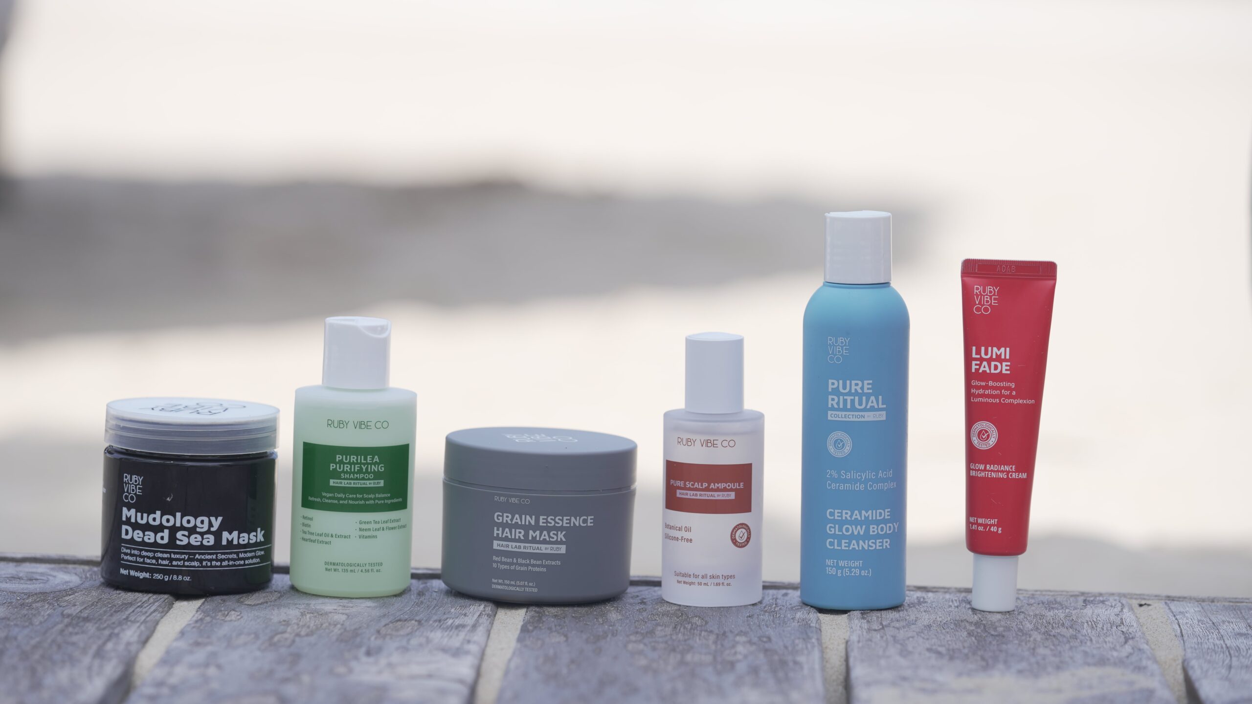 A collection of Ruby Vibe Co. hair and body care products, including the Purilea Purifying Shampoo and Mudology Dead Sea Mask, are arranged on a wooden surface outdoors. The full line of products is shown