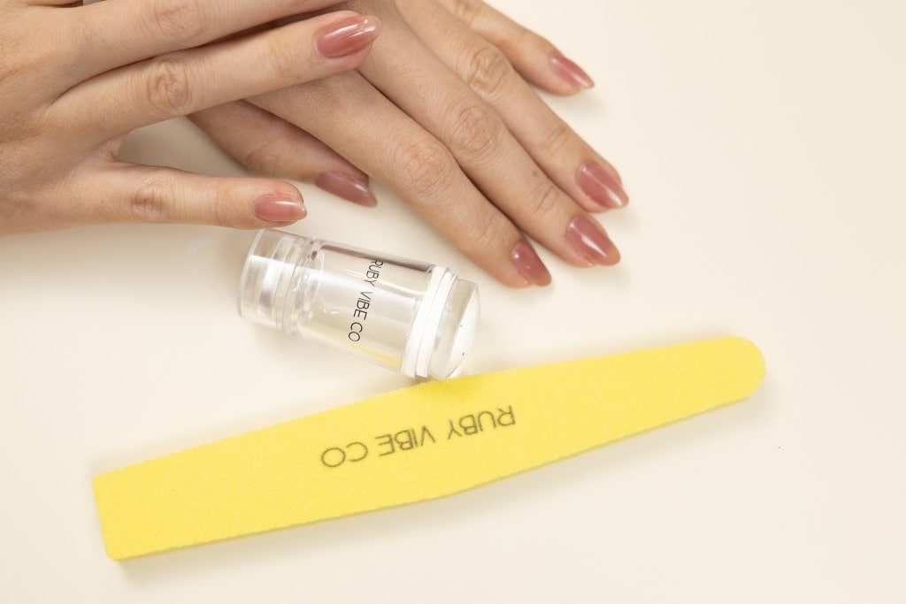 Hands with a glossy, natural-looking manicure displayed next to a yellow Ruby Vibe Co nail file and a clear nail stamper.
