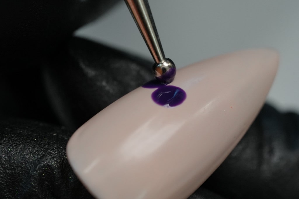 A close-up of a nail dotting tool creating a small, dark purple dot on a nude-colored artificial nail for a nail art design.