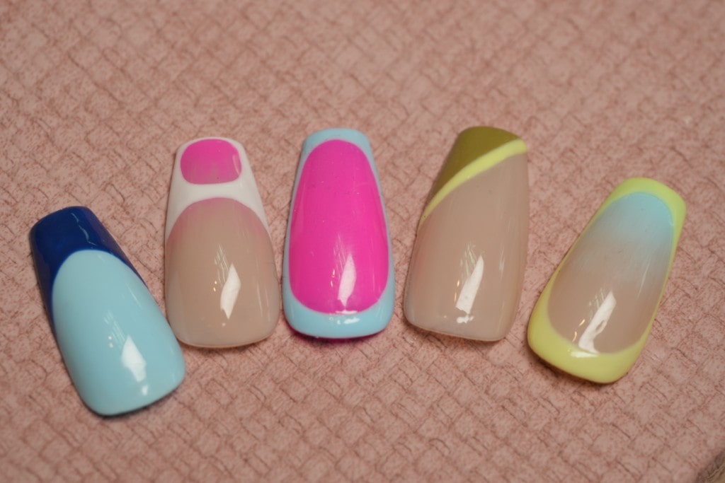  Five artificial nail tips showcasing various modern and colorful gel nail art designs on a pink textured surface.