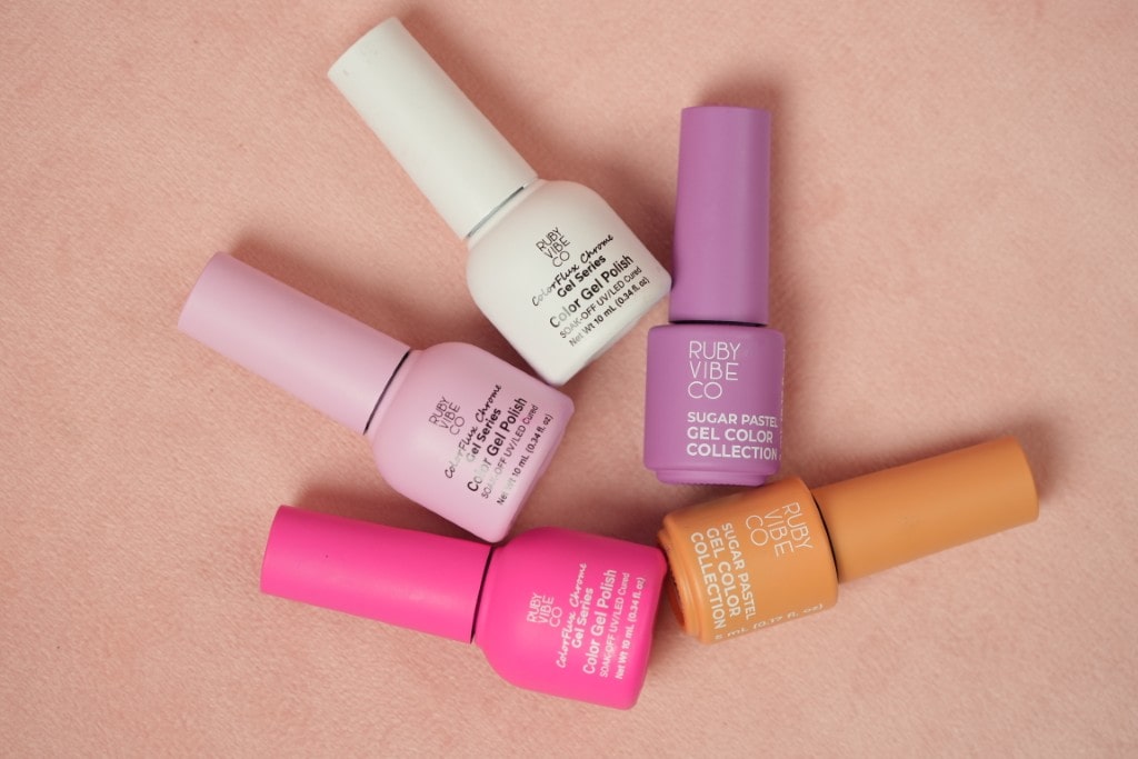 An overhead view of five colorful bottles of Ruby Vibe Co gel polish in shades of pink, purple, white, and orange on a pink background.