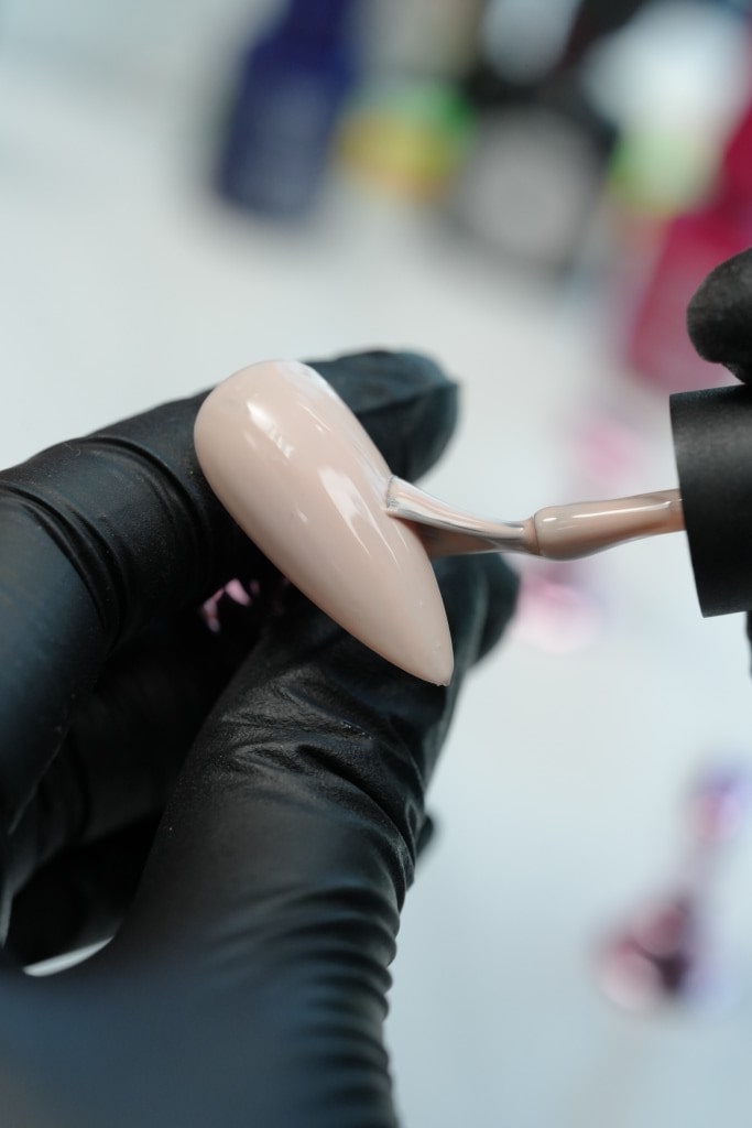 A nail technician in a black glove carefully applies a smooth coat of nude gel polish to a long, almond-shaped nail tip.