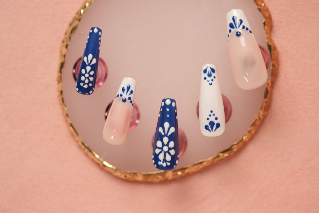  Five long, coffin-shaped press-on nails with intricate blue and white floral designs displayed on a pink surface.