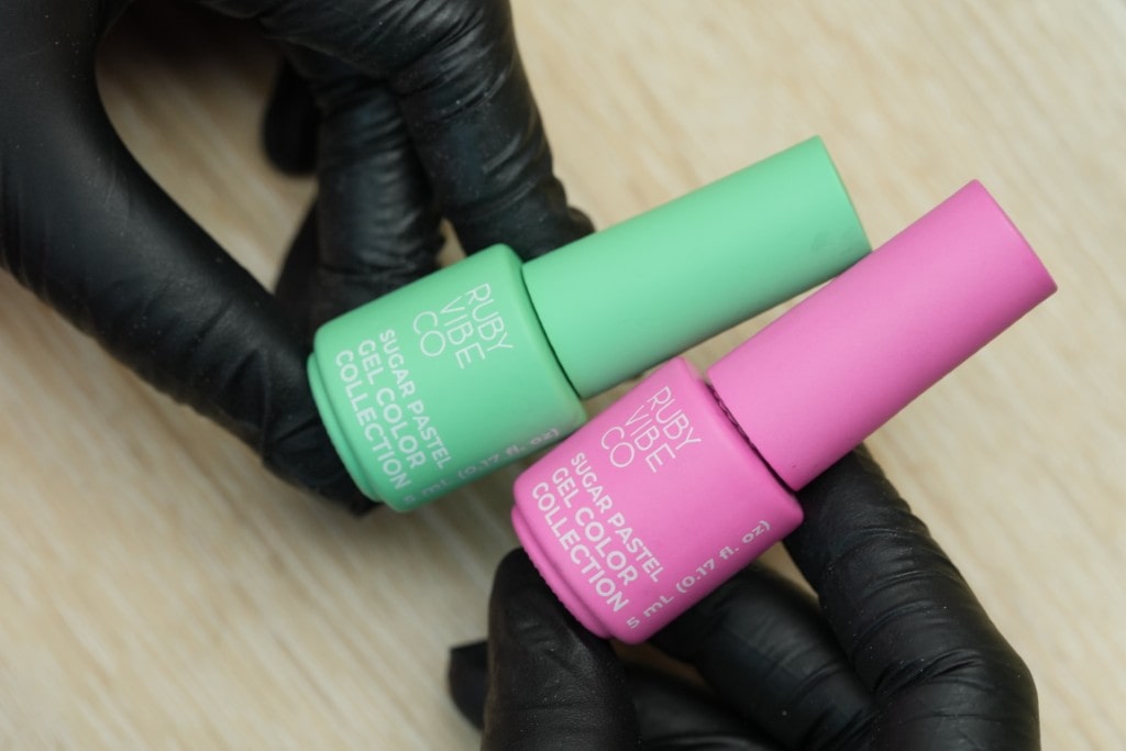 A person wearing black gloves holds up two pastel nail polish bottles, one mint green and one pink, from the Ruby Vibe Co Sugar Pastel Collection.