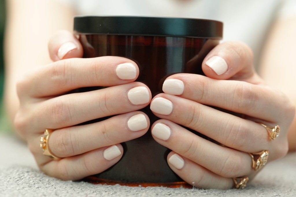  Two hands with a simple, clean manicure in a creamy off-white color are wrapped around a dark brown jar.